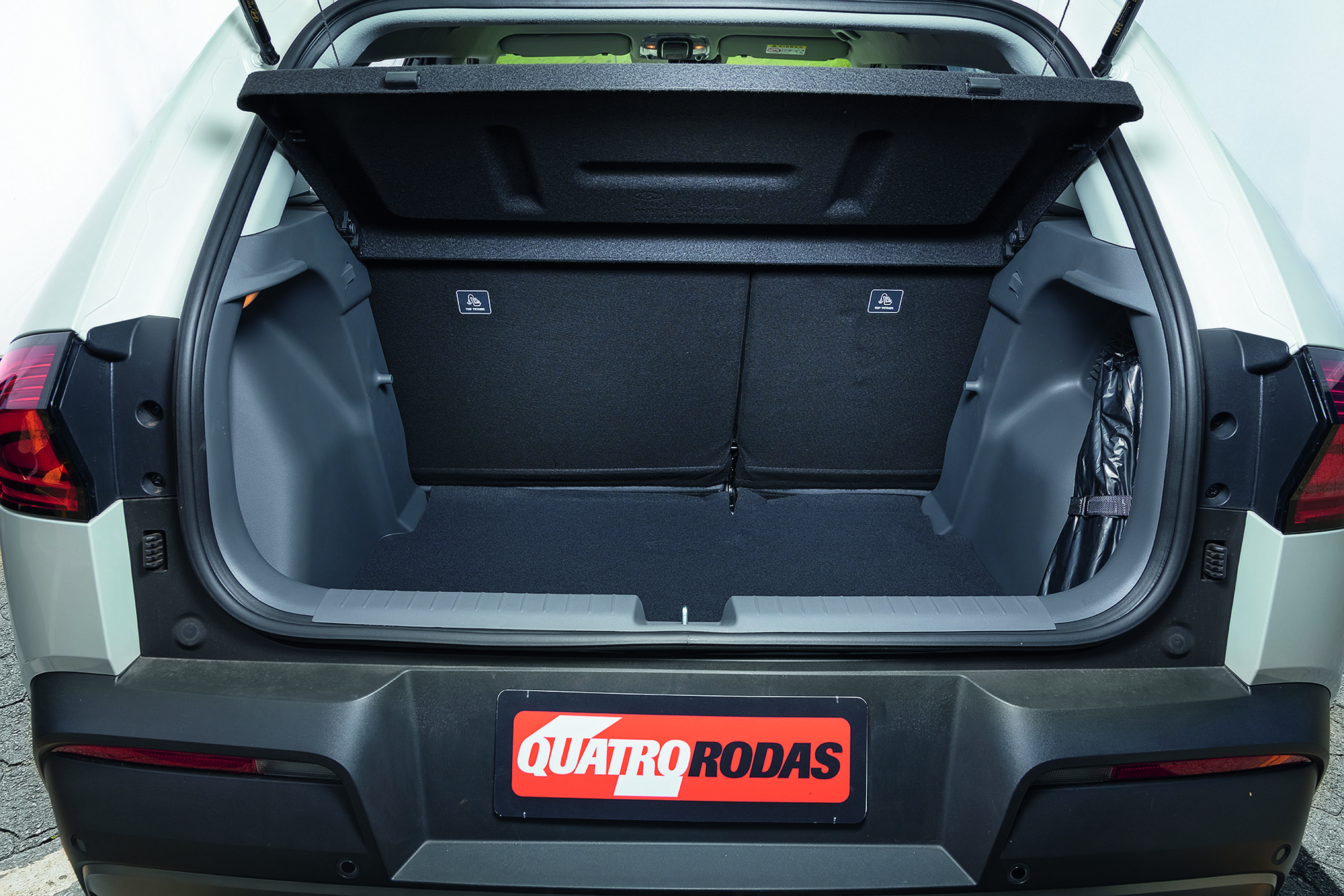 Traseira de um carro branco com o porta-malas aberto, mostrando o interior espaçoso e forrado em preto. Um item preto enrolado está preso na lateral direita. A placa traseira exibe o logotipo QUATRO RODAS em vermelho e branco