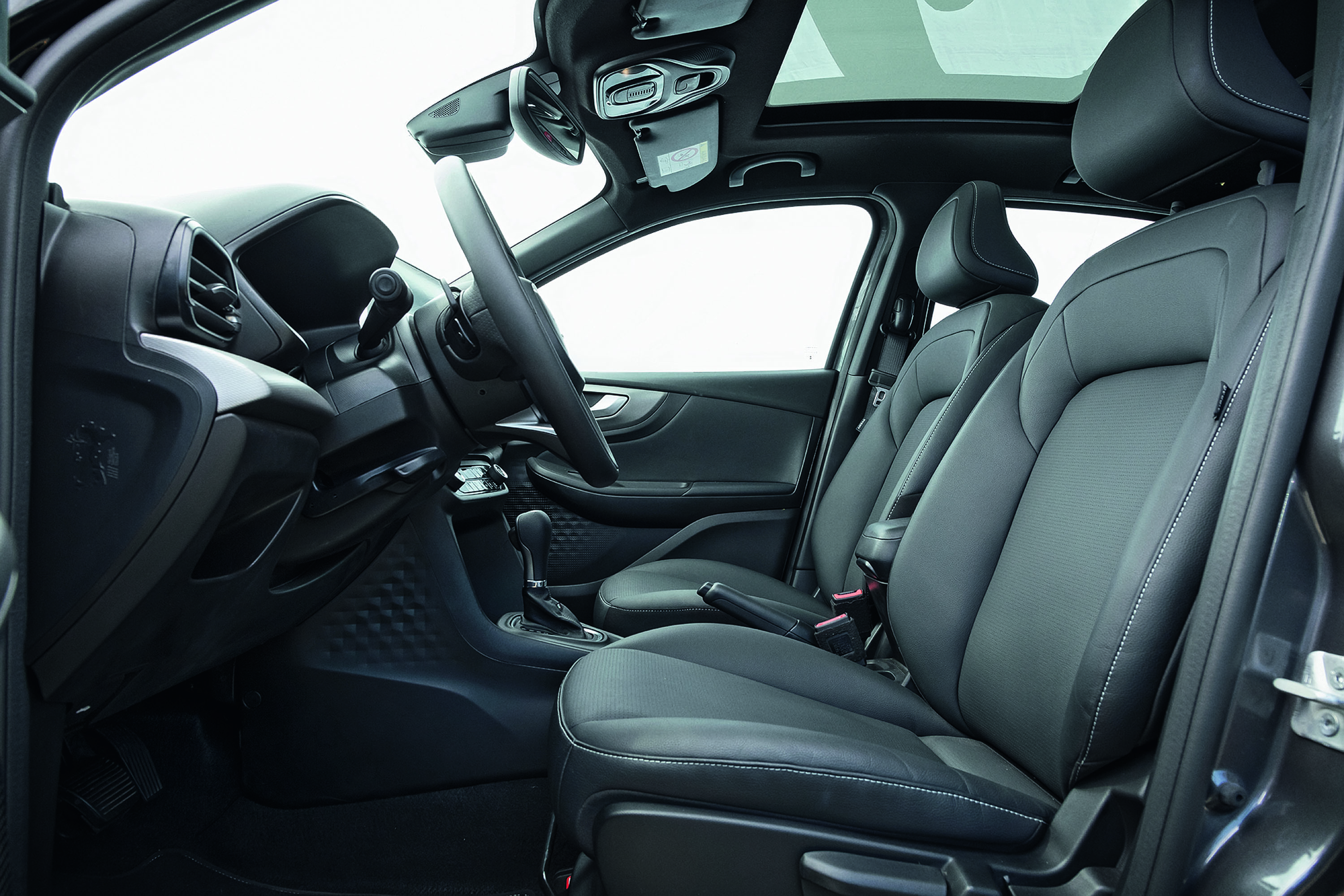 Interior de um carro moderno com bancos de couro escuro e costura branca, volante à esquerda, câmbio automático e teto solar panorâmico. O painel e as portas são pretos, com detalhes texturizados. As janelas mostram um fundo branco, sugerindo um estúdio.