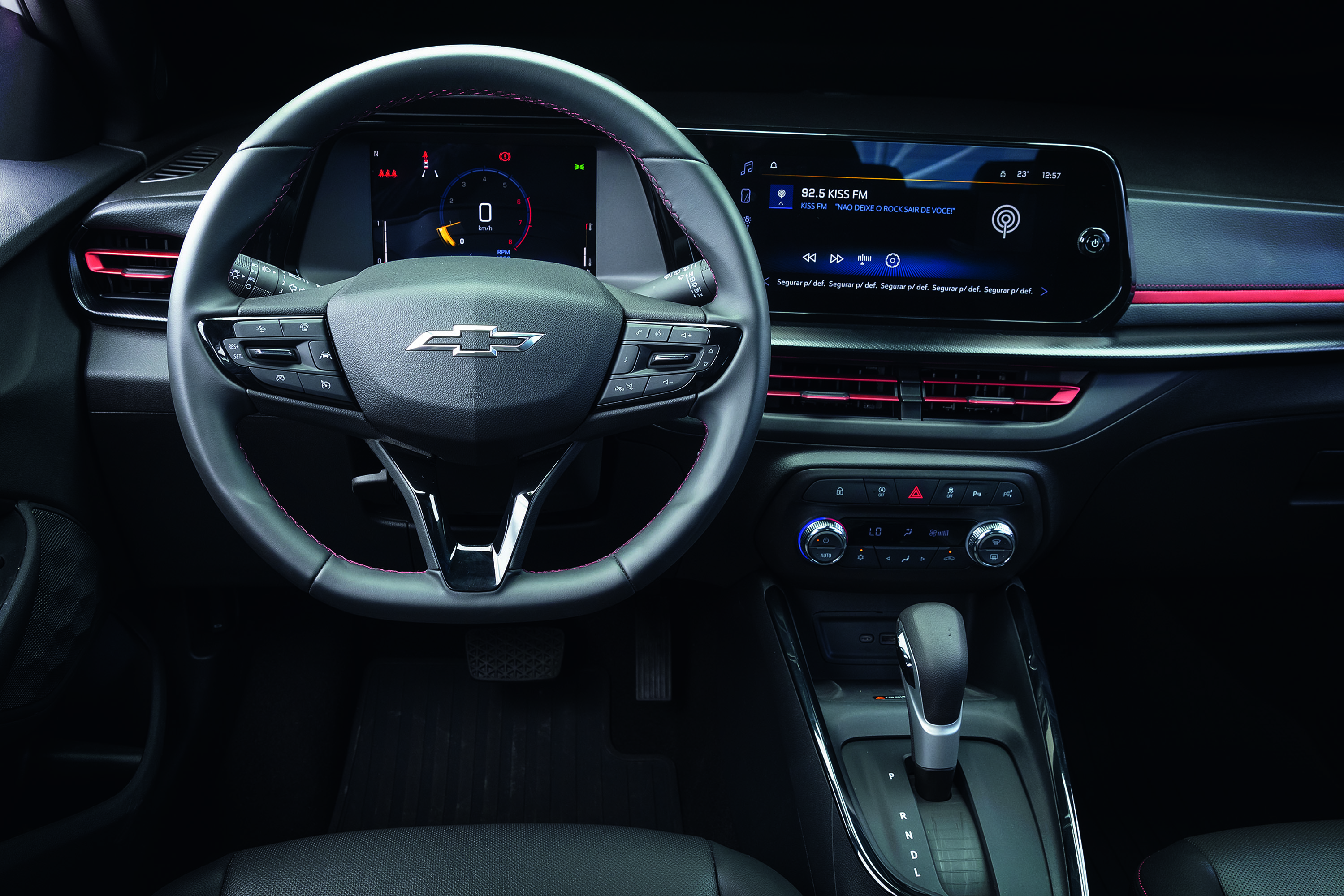 Interior de um carro moderno com volante multifuncional da Chevrolet em primeiro plano, painel digital com velocímetro marcando zero e tela multimídia exibindo rádio 92.5 Kiss FM. Detalhes em vermelho iluminam as saídas de ar e o painel, com câmbio automático e botões de controle de ar-condicionado visíveis abaixo