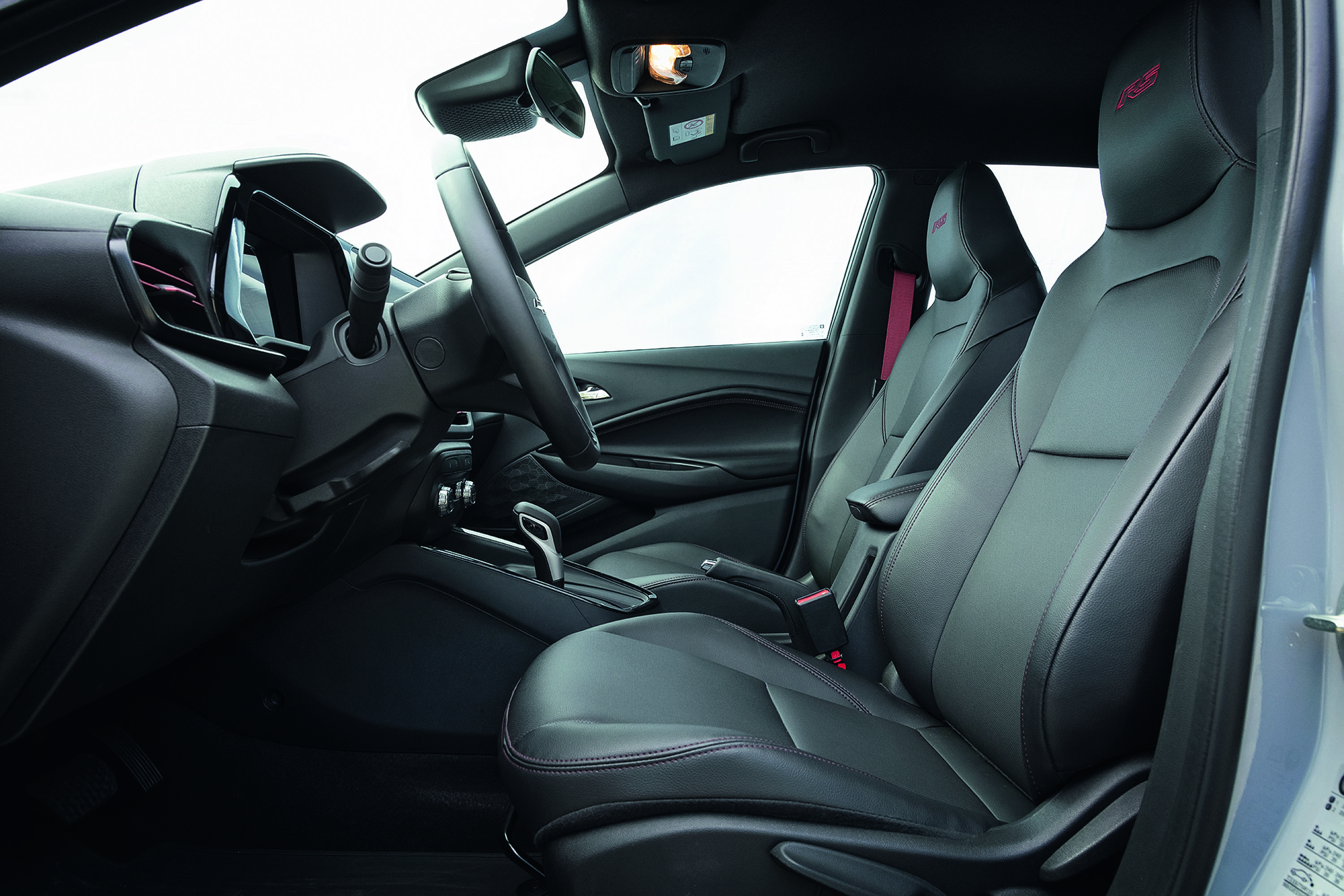 Interior de um carro com bancos de couro pretos e detalhes vermelhos. O volante e o painel são escuros, com uma tela digital visível. O cinto de segurança do passageiro é vermelho, contrastando com o estofamento. A alavanca de câmbio automática está no console central.