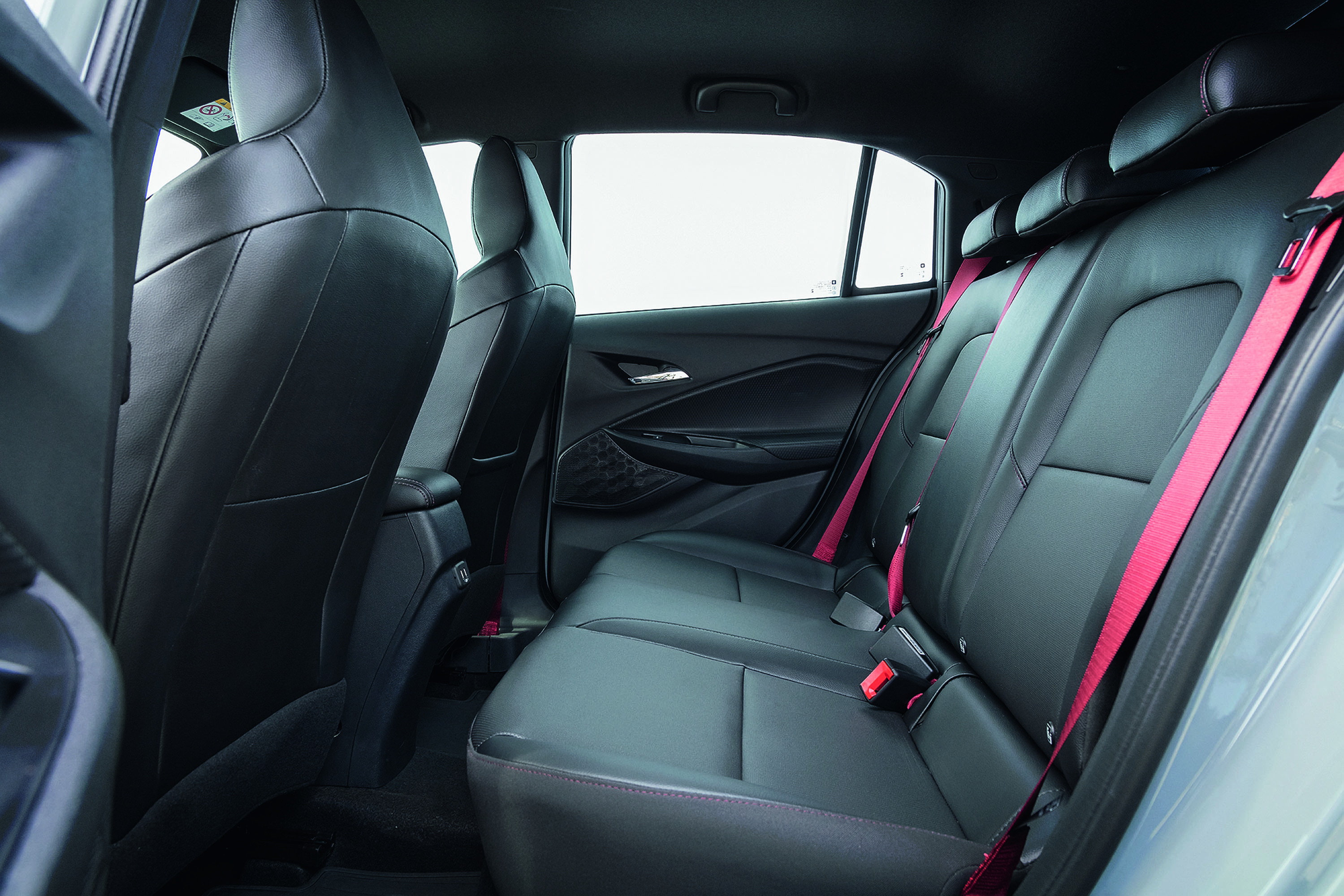 Interior de um carro com bancos traseiros de couro preto e costura vermelha, cintos de segurança vermelhos vibrantes, e as portas abertas revelando o design interno