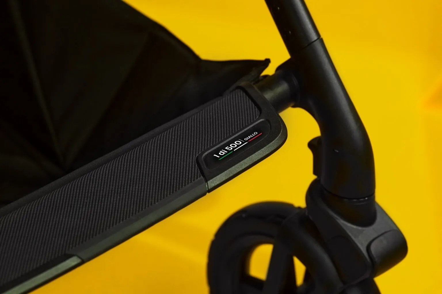 Detalhe de um carrinho de bebê preto com o texto 1 di 500 Giallo e uma pequena bandeira italiana, sobre um fundo amarelo vibrante