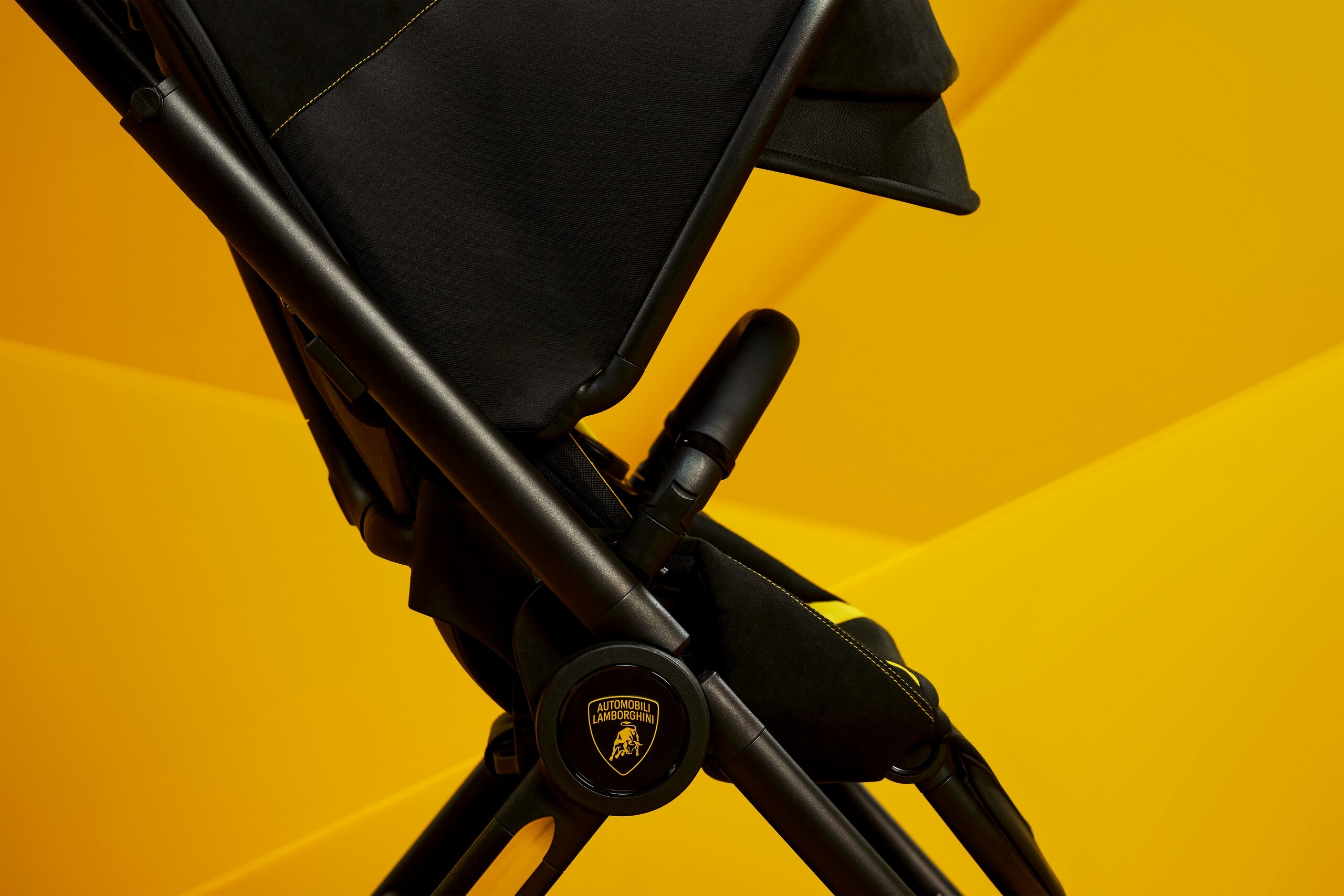 Carrinho de bebê preto com detalhes amarelos, incluindo o logotipo dourado da Automobili Lamborghini em uma roda, contra um fundo amarelo vibrante