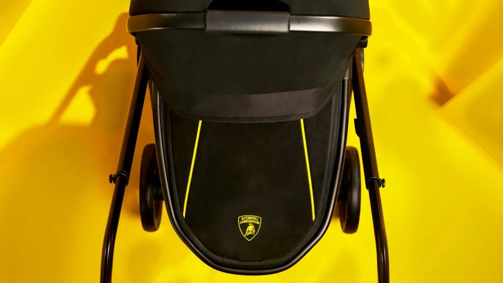 Carrinho de bebê preto com detalhes amarelos, incluindo duas linhas verticais e o logotipo da Lamborghini, em fundo amarelo vibrante