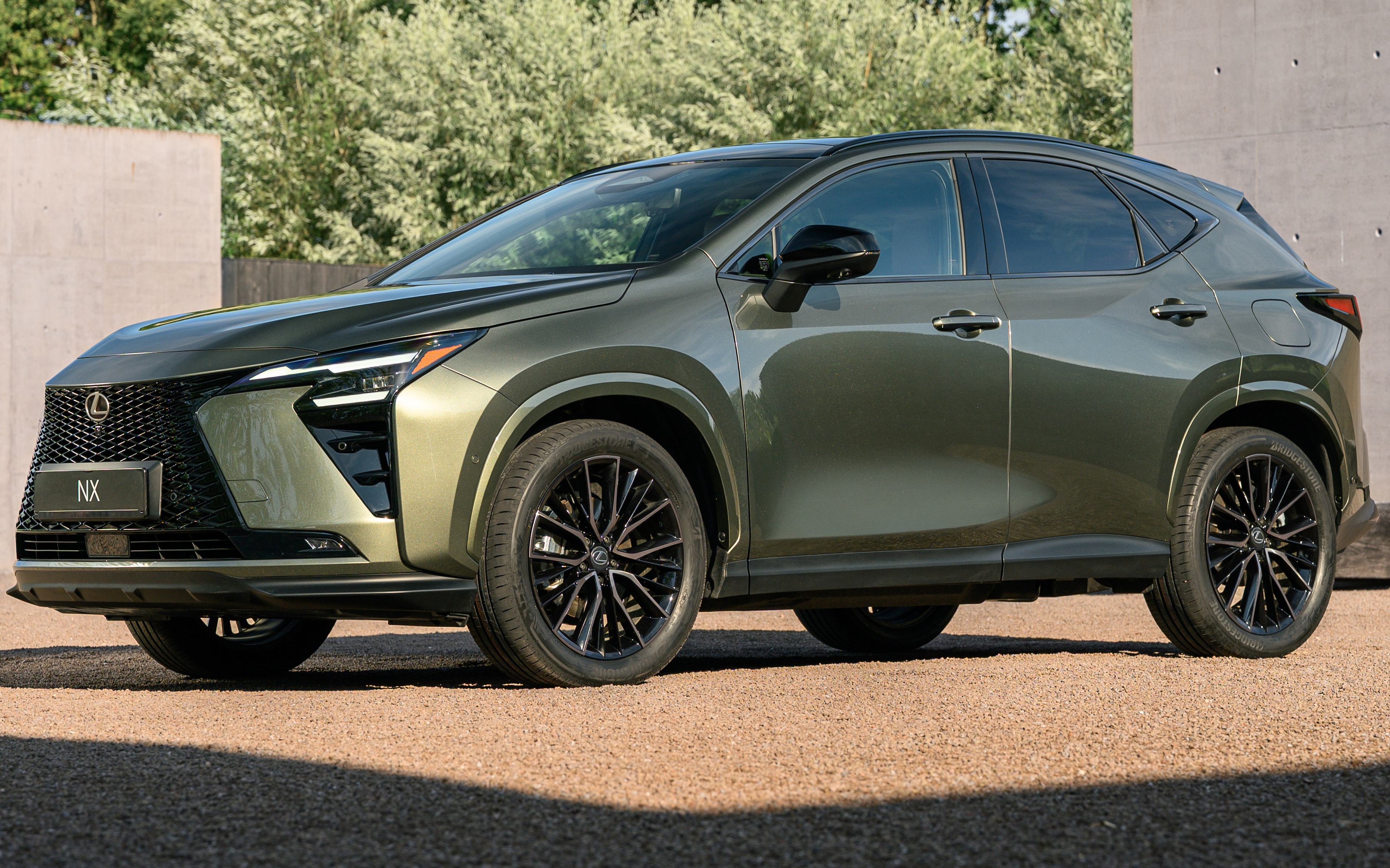 SUV Lexus NX 2024 verde-oliva metálico, estacionado em uma área externa com piso de cascalho e vegetação ao fundo. O carro tem rodas pretas e detalhes cromados na grade frontal e faróis. A luz do sol incide sobre o veículo, criando sombras no chão