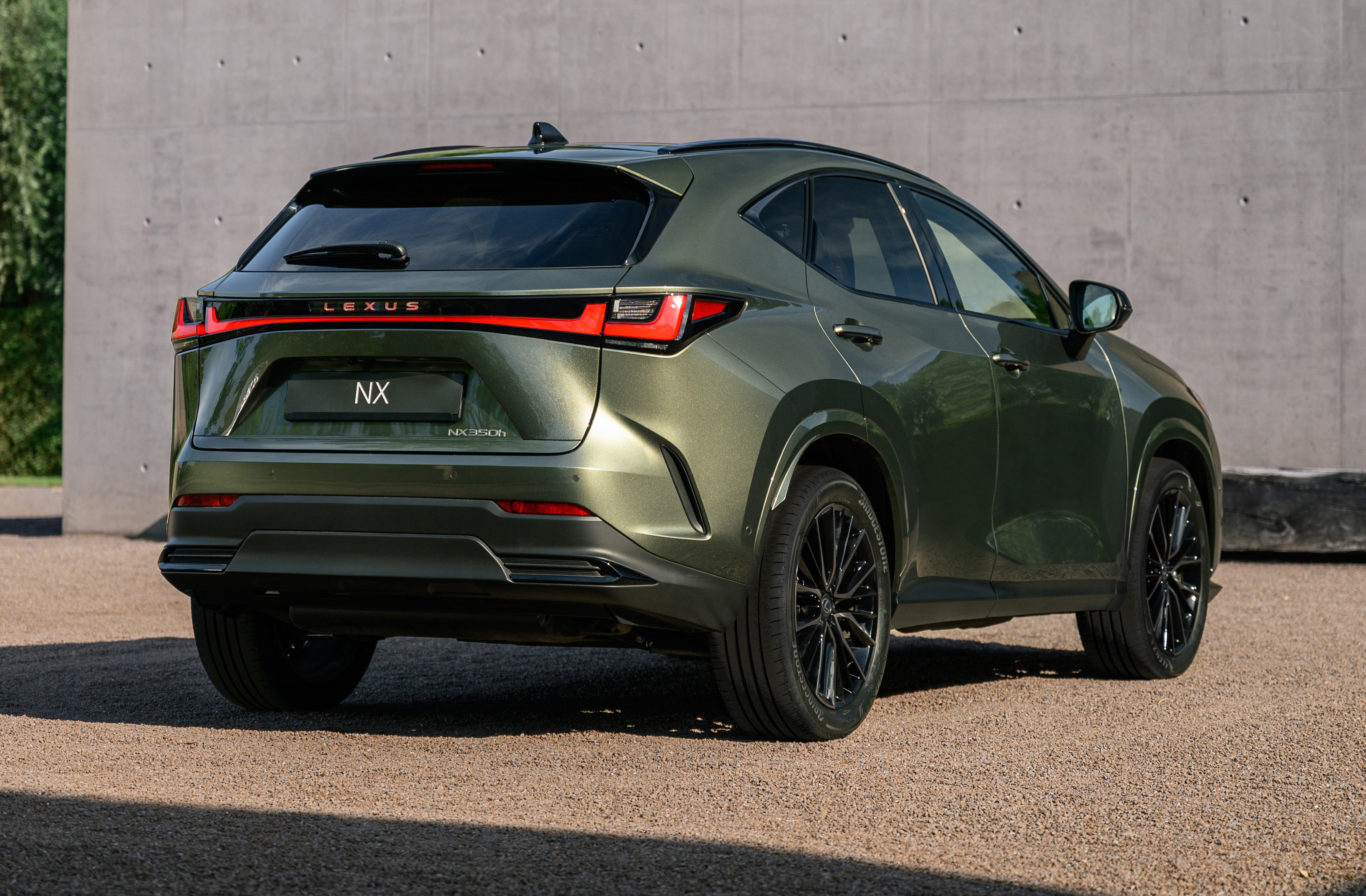 Traseira de um SUV Lexus NX verde-oliva metálico, estacionado em uma superfície de cascalho. As lanternas traseiras em LED formam uma linha horizontal vermelha, com o nome LEXUS acima e NX no centro da tampa do porta-malas. Rodas pretas com múltiplos raios e detalhes cromados. Ao fundo, uma parede de concreto cinza e vegetação verde à esquerda