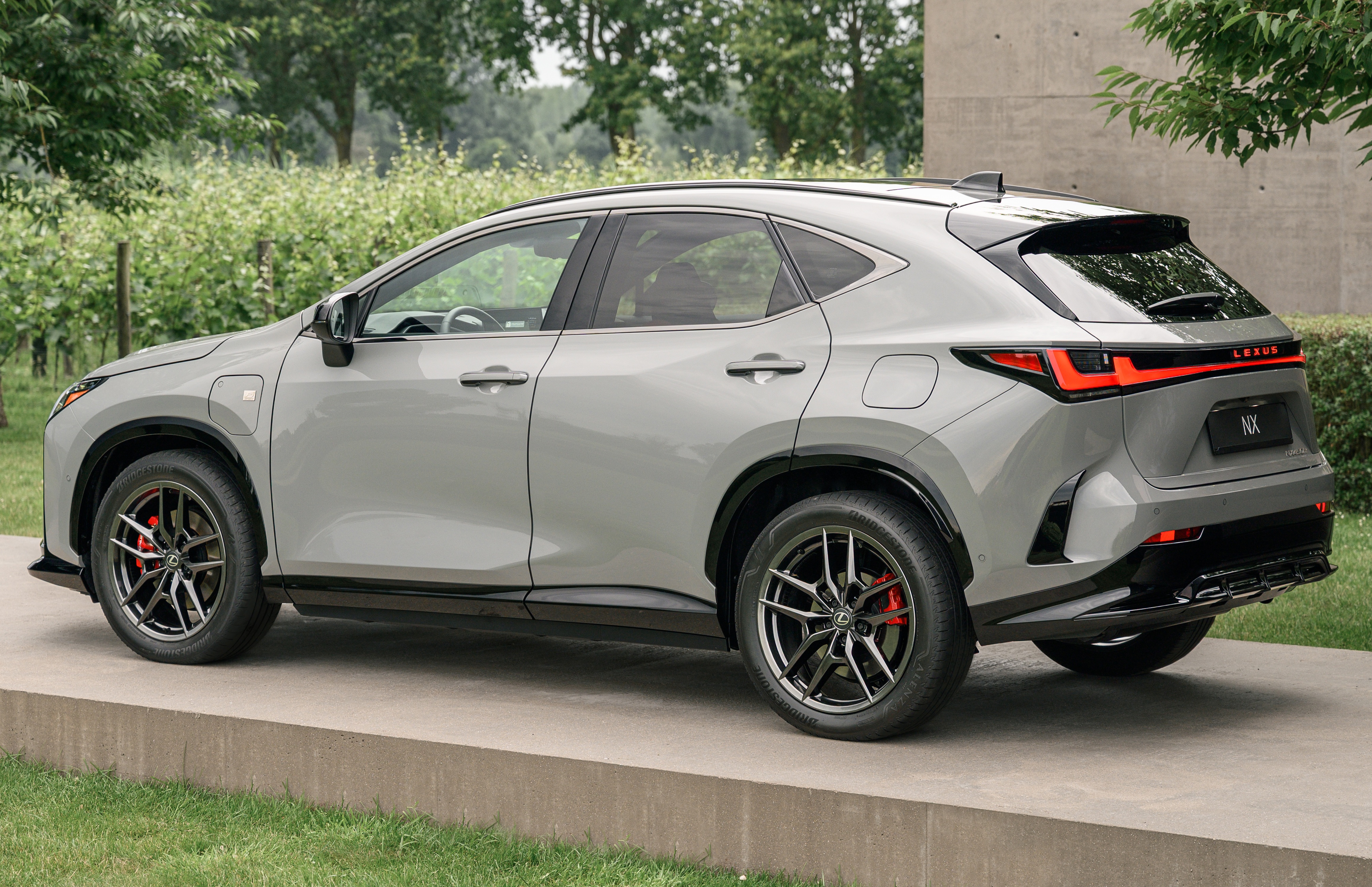 SUV Lexus NX cinza claro, visto de lado, estacionado em uma plataforma de concreto com grama ao redor. O carro tem detalhes pretos nas rodas e na parte inferior da carroceria, e lanternas traseiras em LED vermelhas e pretas. Ao fundo, árvores e vegetação verde