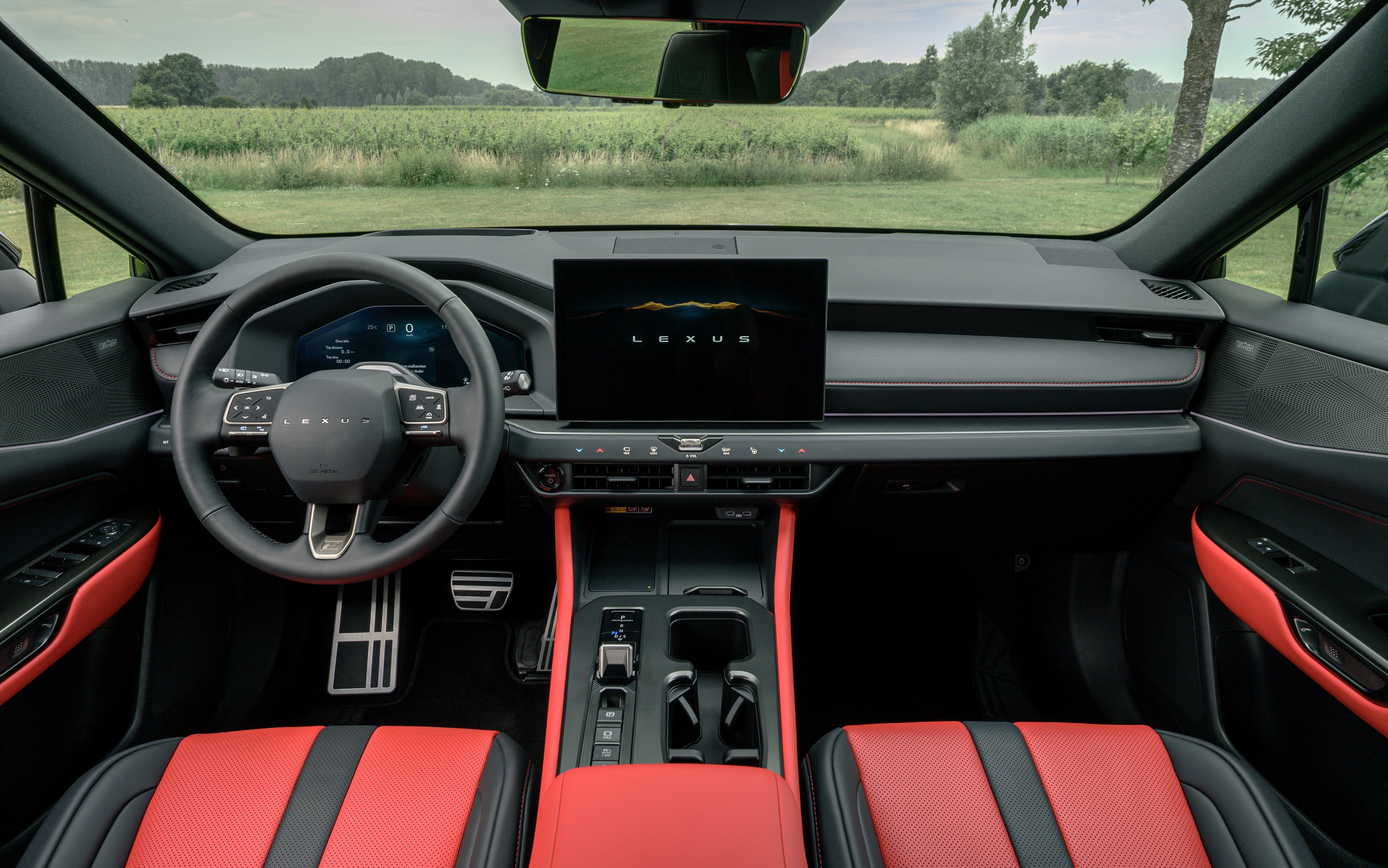 Interior de um carro Lexus com bancos esportivos vermelhos e pretos, volante multifuncional escuro e painel com tela central exibindo o logo Lexus. O para-brisa revela um campo verde com árvores ao fundo sob um céu claro