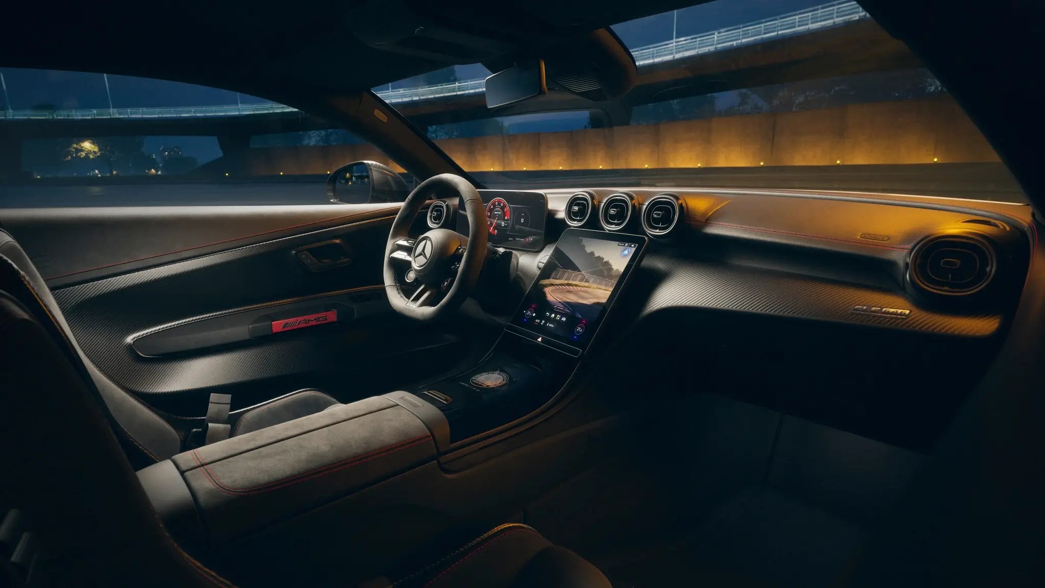 Interior de um carro esportivo Mercedes-Benz à noite, com painel escuro de fibra de carbono, volante com o logo da marca, tela digital central exibindo um cenário de estrada e luzes laranja refletindo no para-brisa. O banco do motorista, parcialmente visível, é de camurça escura com costuras vermelhas