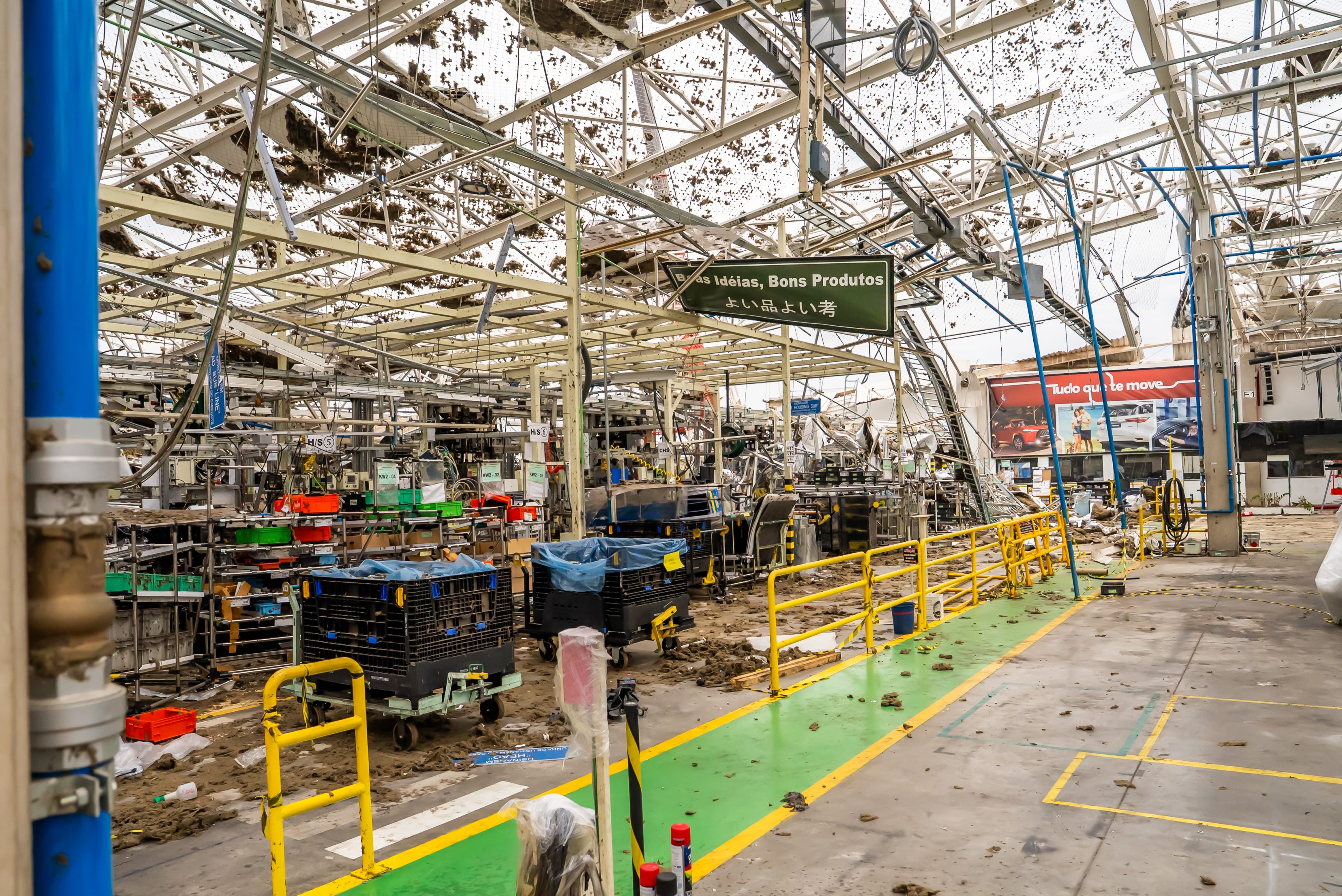Interior de uma fábrica destruída, com o teto desabado e estruturas metálicas retorcidas. O chão está coberto por detritos e lama, com carrinhos de transporte e equipamentos danificados. Uma placa verde pendurada exibe Boas Ideias, Bons Produtos em português e japonês. Ao fundo, um outdoor vermelho com Tudo que se move e imagens de carros. O ambiente é de devastação pós-desastre.