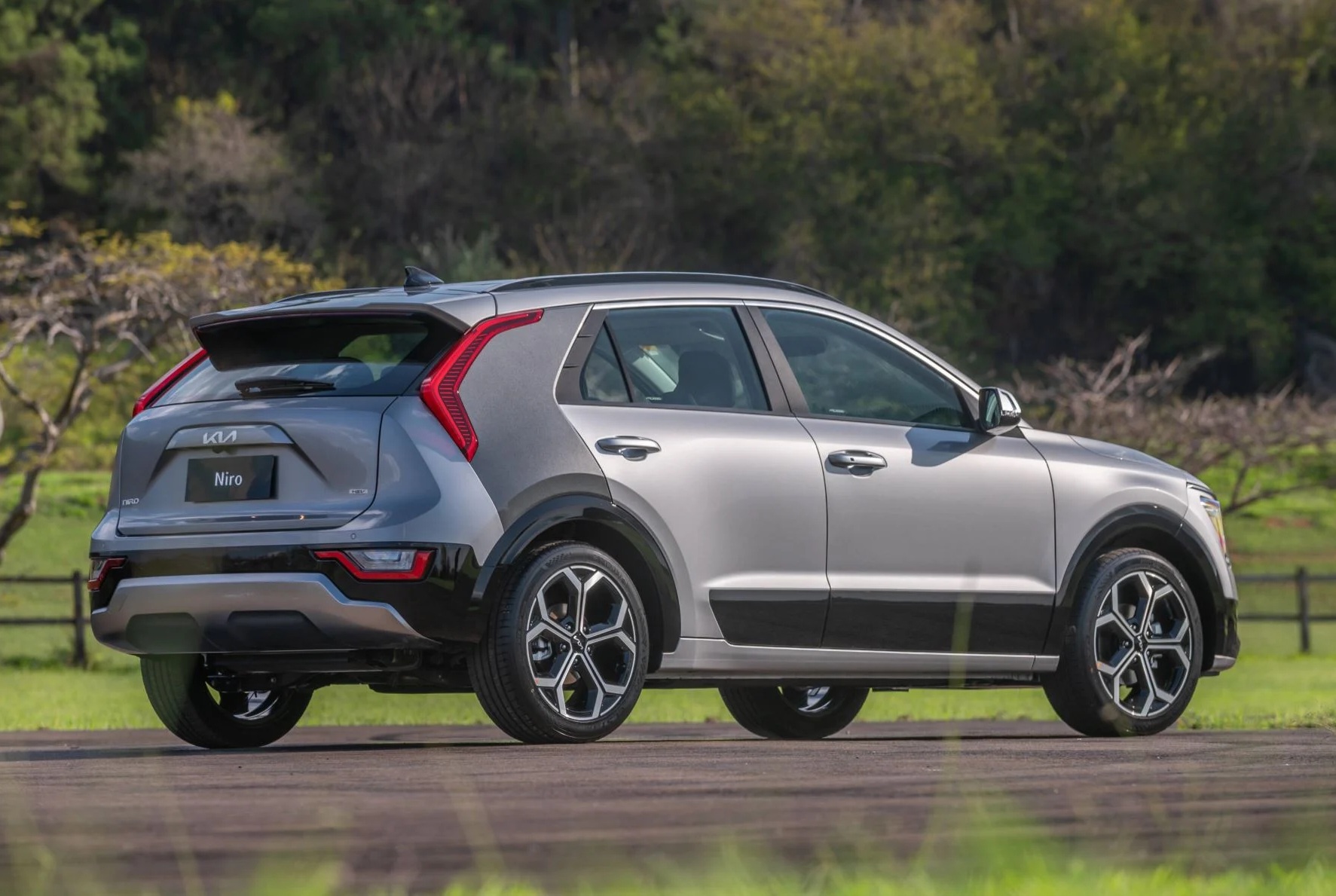 SUV Kia Niro cinza claro e escuro, visto de traseira e lateral, estacionado em uma estrada asfaltada com grama e árvores ao fundo