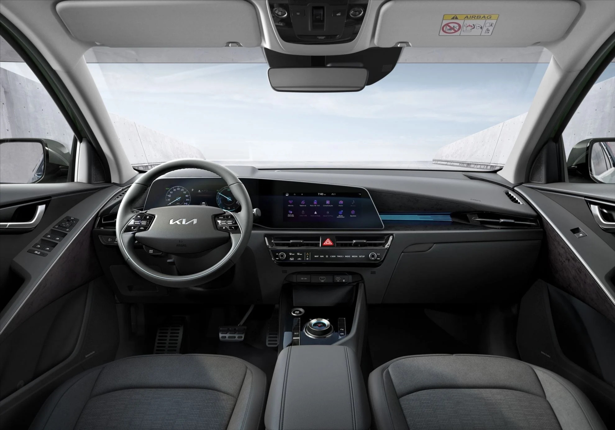 Interior de um carro moderno com painel cinza escuro, volante multifuncional com logo Kia, tela multimídia central e painel de instrumentos digital. Os bancos são de tecido cinza e o para-brisa mostra uma estrada com céu claro