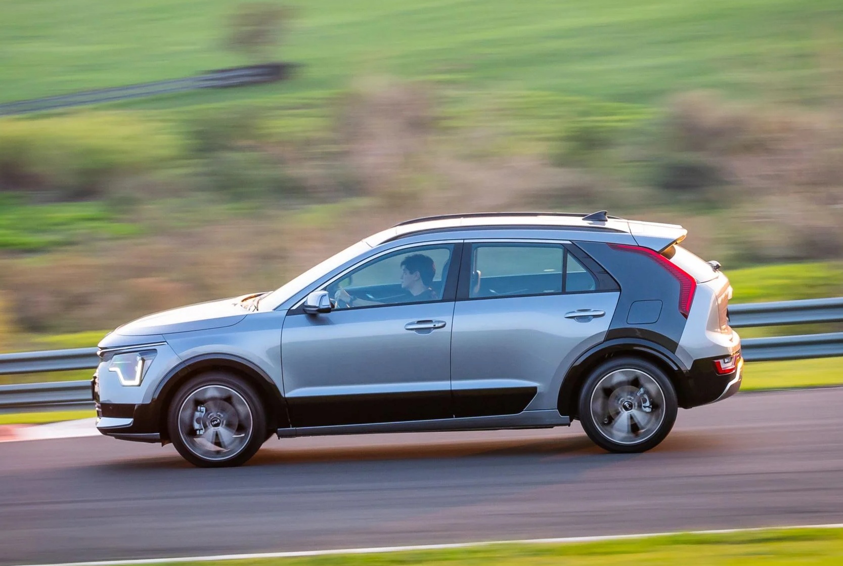 Carro SUV prateado com detalhes pretos na parte inferior, em movimento rápido em uma pista de corrida, com um motorista visível ao volante. O fundo verde está borrado, indicando velocidade
