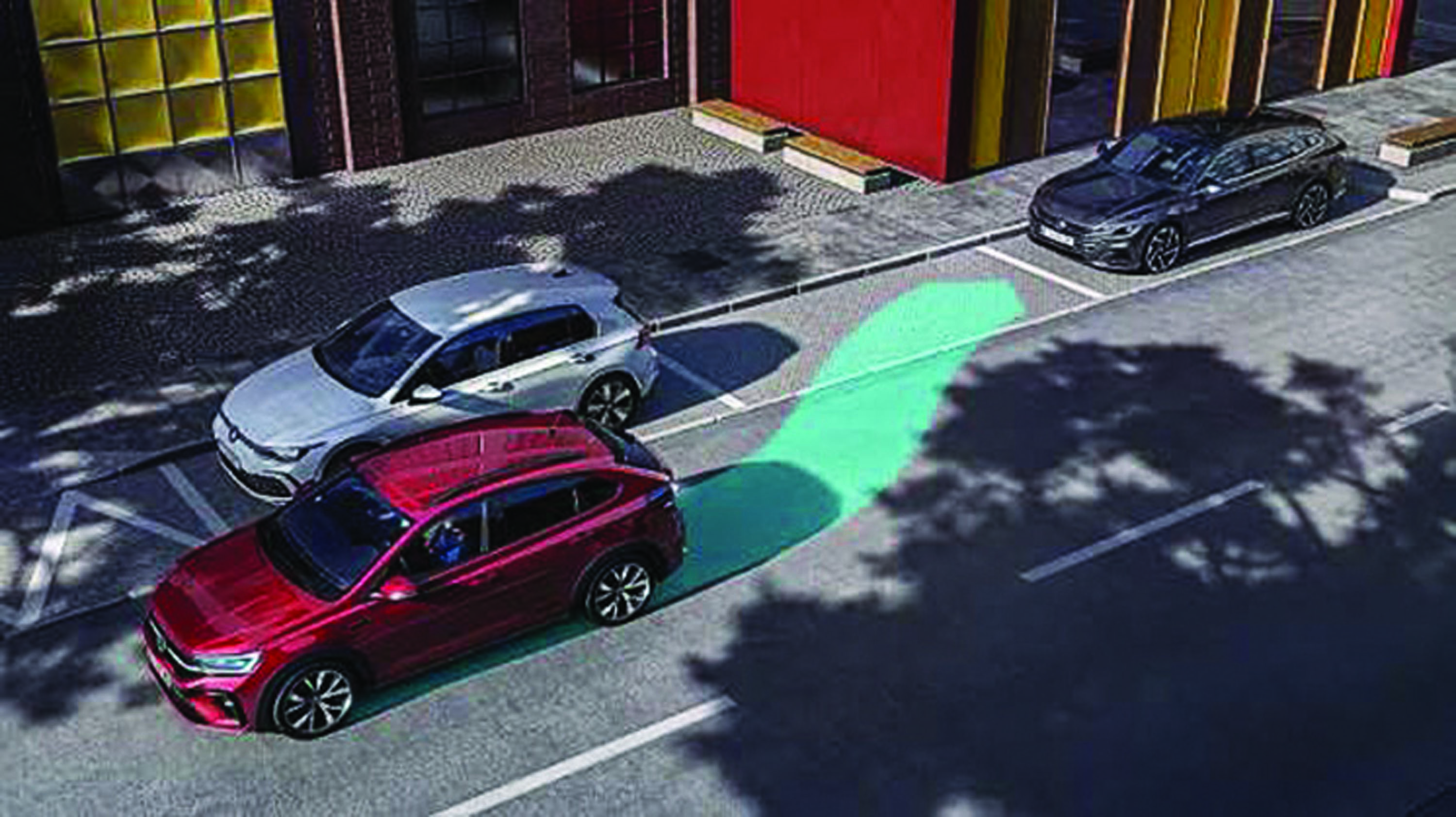Vista aérea de três carros estacionados em uma rua. Um carro vermelho está em primeiro plano, um prata atrás dele e um preto mais distante. Uma faixa verde brilhante no asfalto indica uma vaga de estacionamento. Sombras de árvores e edifícios coloridos completam a cena urbana