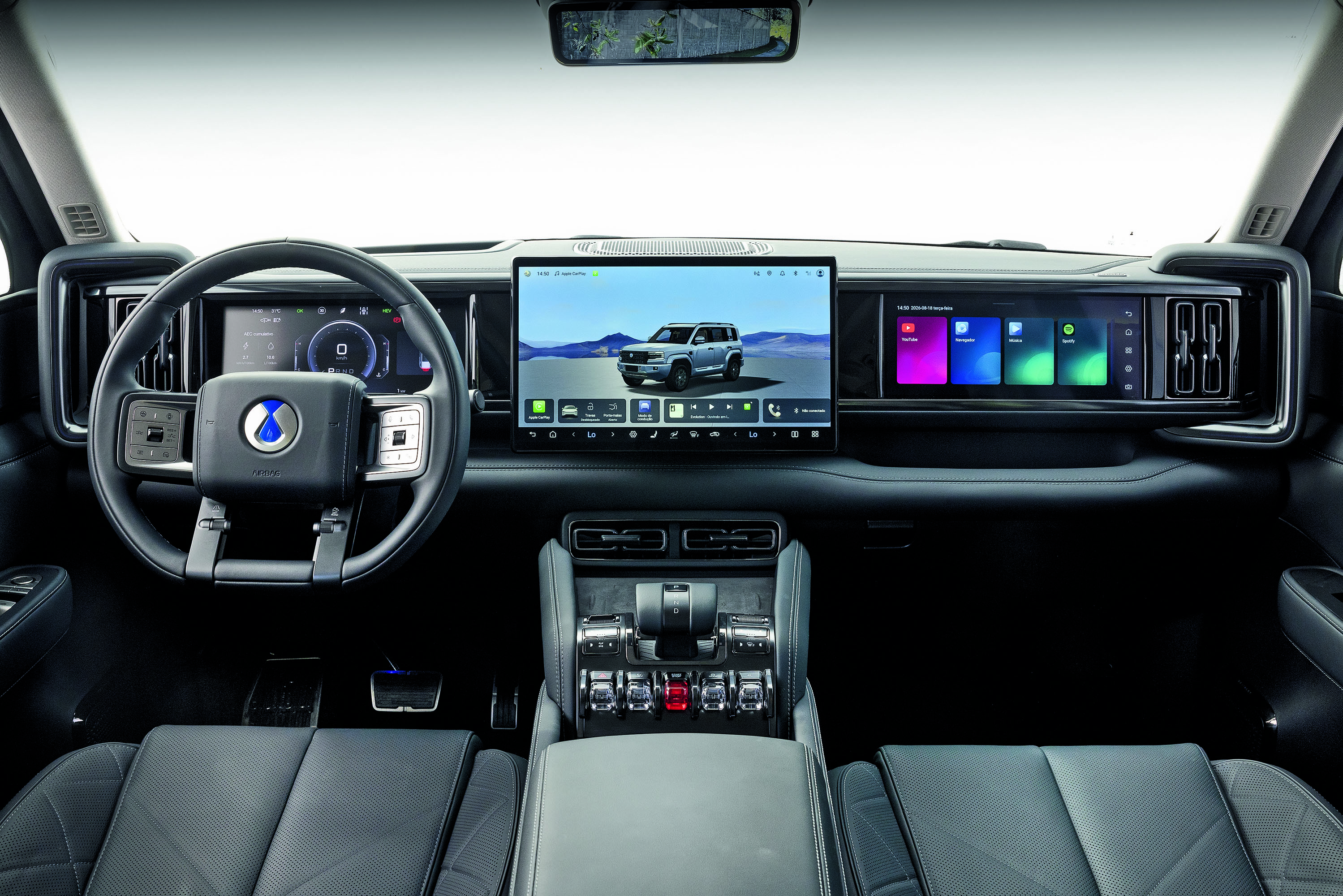Interior de um carro moderno com painel digital. O volante preto tem um logo azul no centro. À direita, uma tela exibe um SUV cinza em paisagem montanhosa. Outra tela menor mostra aplicativos coloridos. Bancos de couro pretos e console central com botões prateados completam o design.