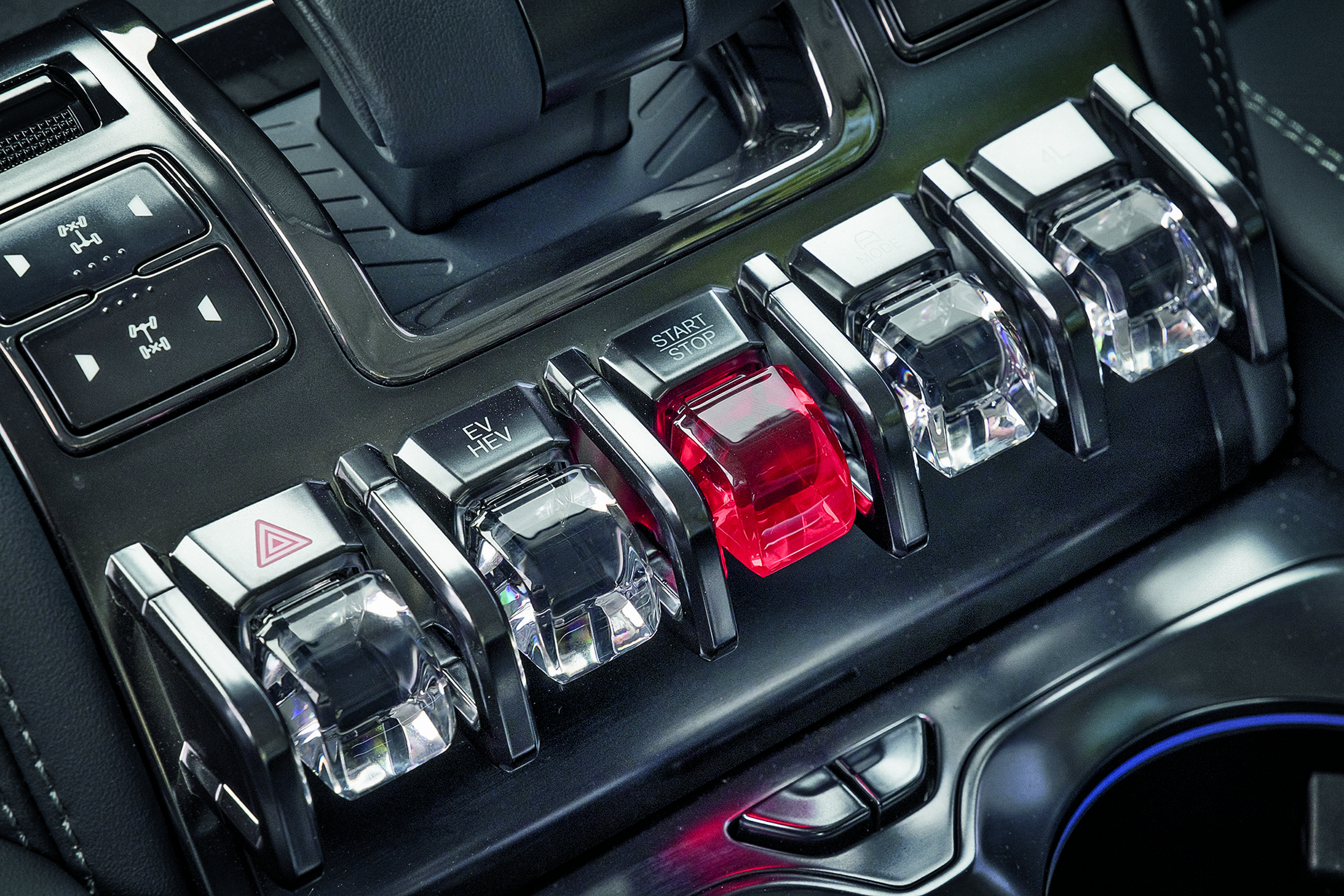 Painel de controle de carro com cinco botões tipo alavanca, o central vermelho com START STOP, e os outros transparentes com símbolos de alerta, tração e modo elétrico