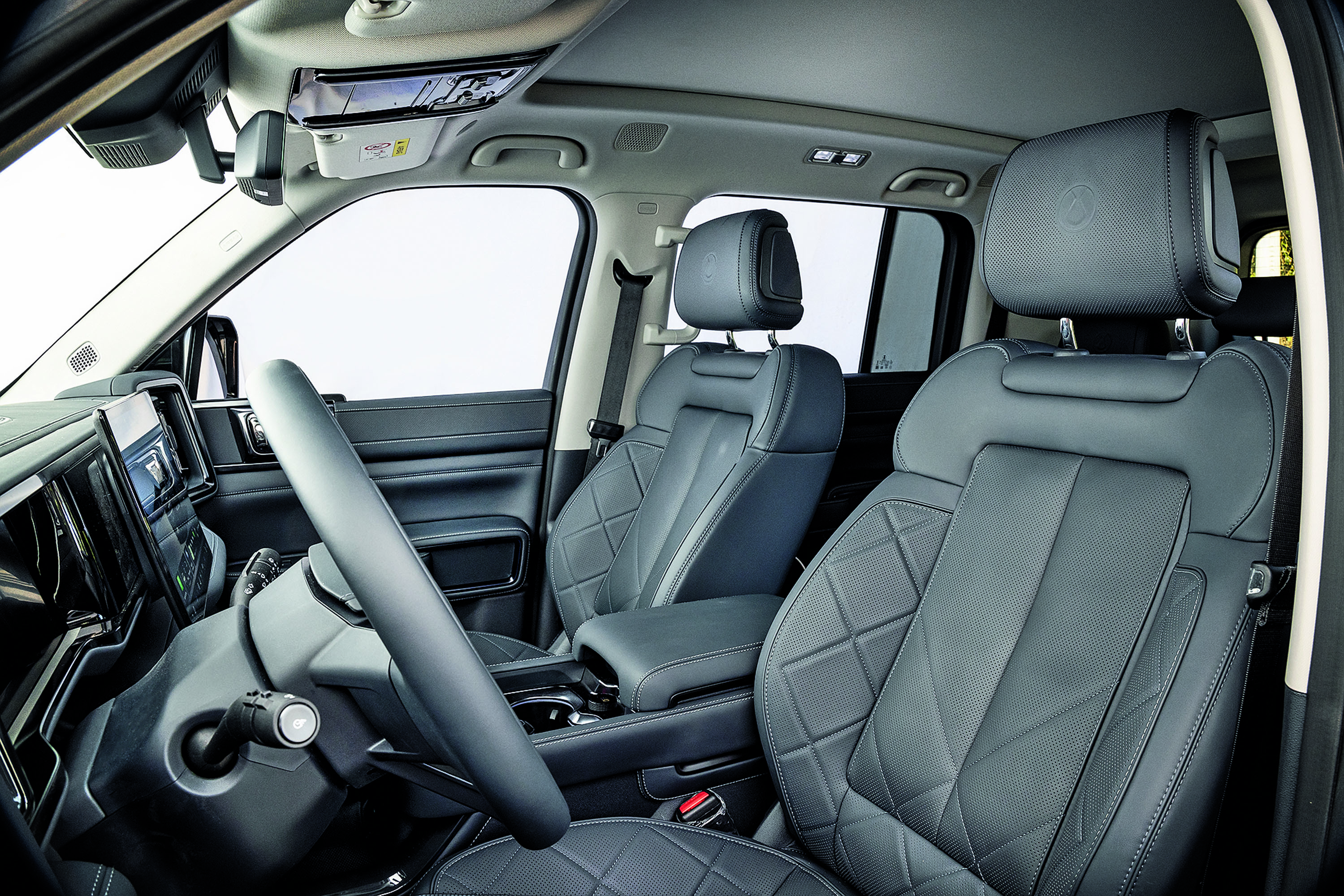 Interior de um carro com bancos de couro cinza escuro e costuras em losango, volante à esquerda e painel digital. O banco do motorista e o do passageiro dianteiro são visíveis, com o banco traseiro parcialmente à direita. O teto solar e o espelho retrovisor interno também aparecem