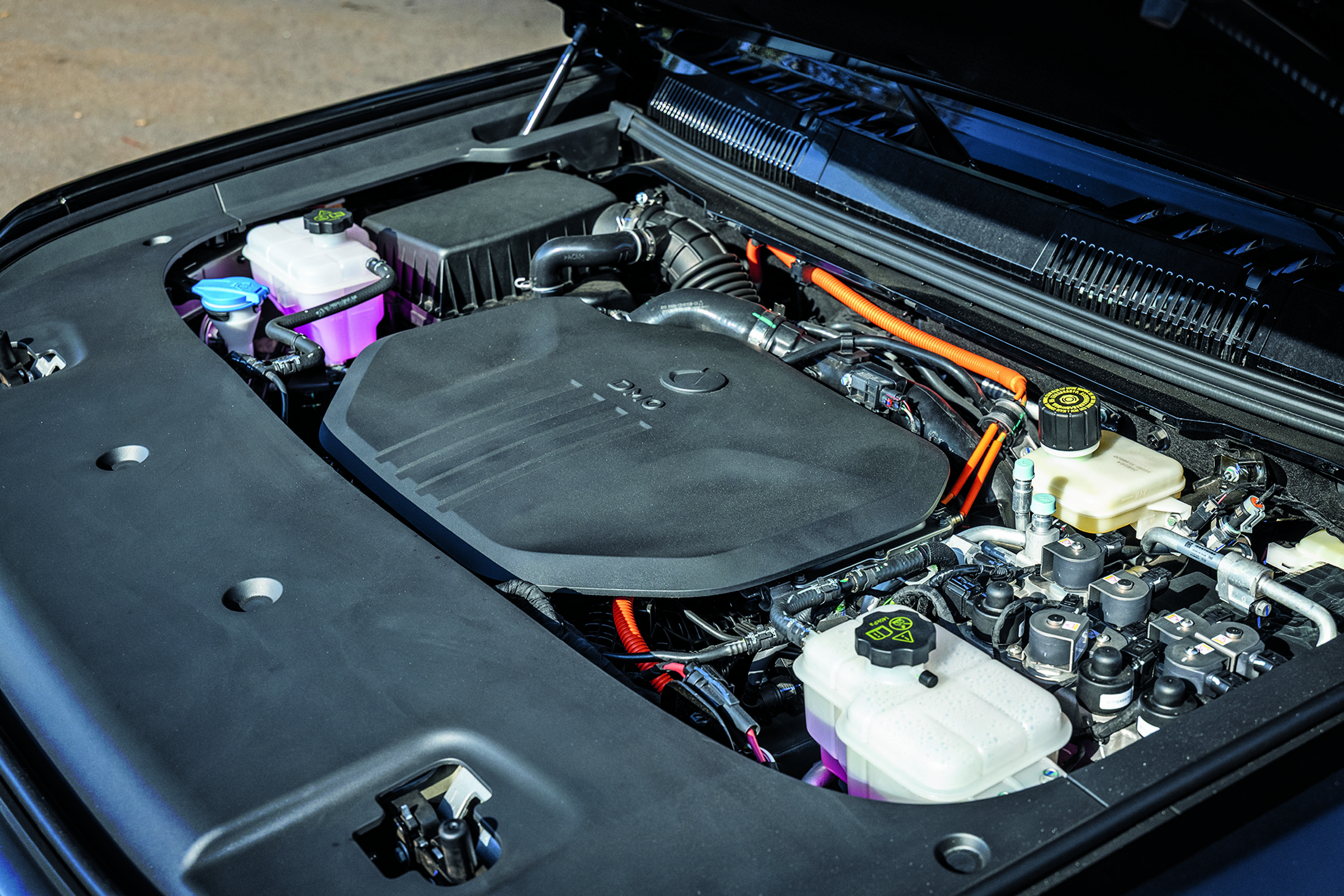Compartimento do motor de um carro híbrido ou elétrico, com cabos laranja de alta voltagem, reservatórios de fluidos e uma tampa preta cobrindo parte do motor