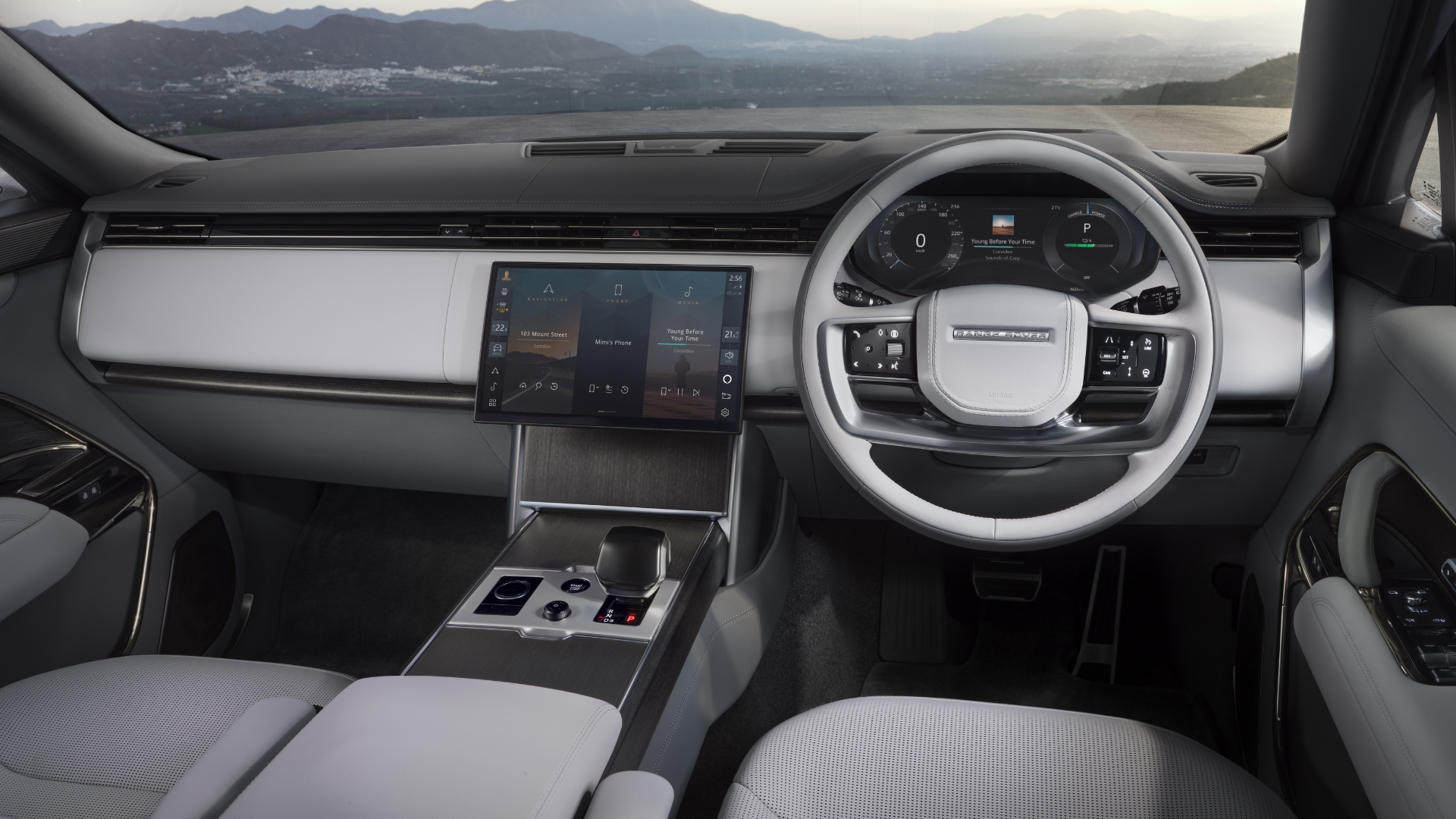 Interior moderno de um carro, com volante branco e painel digital. Uma tela central exibe um menu de mídia, e o console tem acabamento em madeira escura com botões prateados. Bancos claros e montanhas ao fundo completam a cena