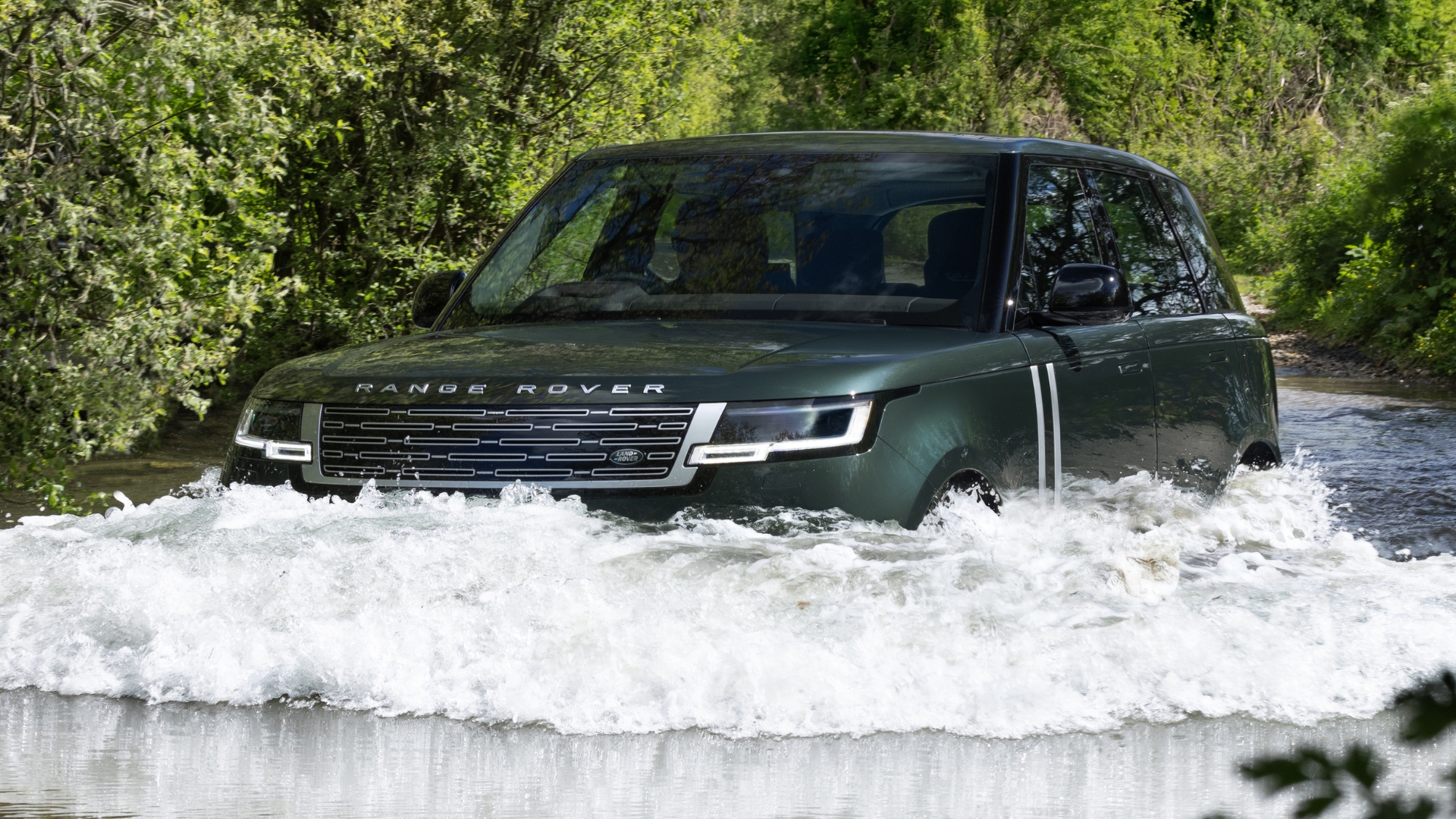 Um SUV Range Rover verde escuro atravessando um rio raso, com água espirrando ao redor dos pneus e da frente do veículo. O carro está em movimento, com árvores verdes ao fundo sob um céu claro