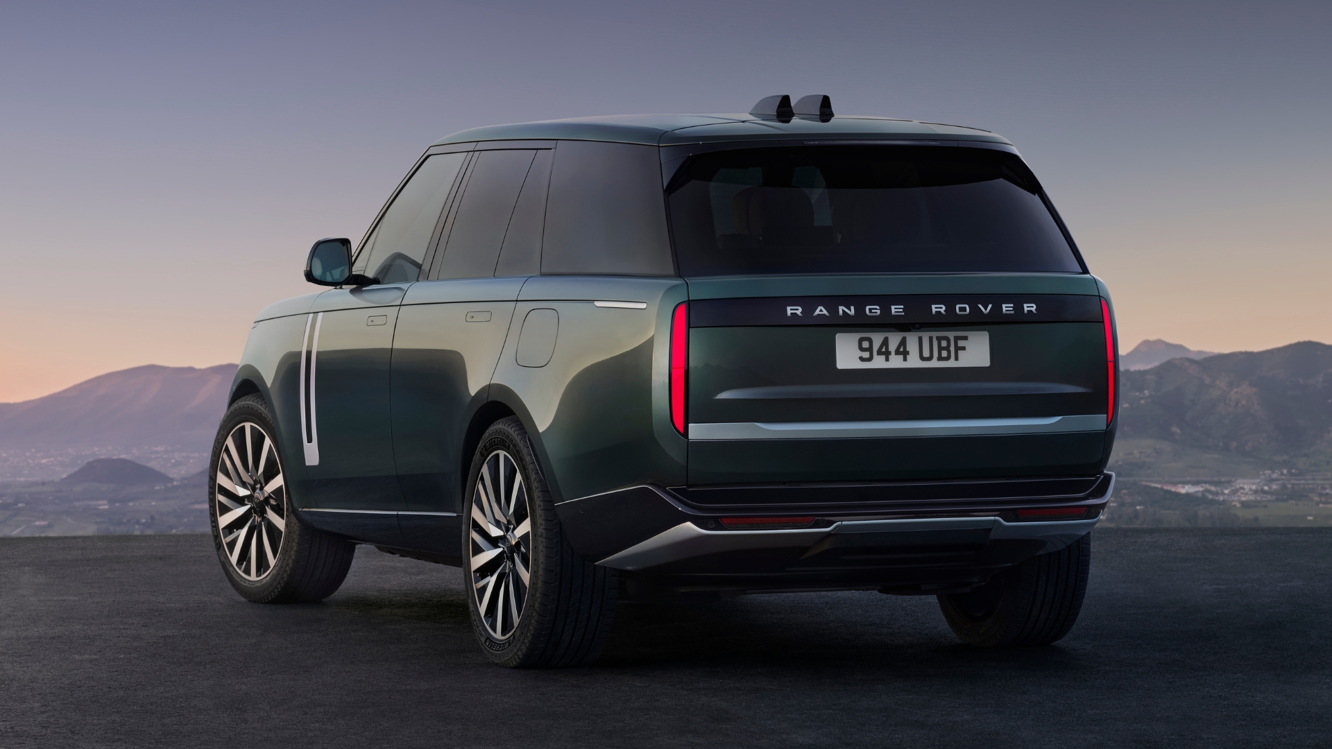 SUV Range Rover verde escuro, visto de traseira e lateral, estacionado em asfalto com montanhas ao fundo sob céu crepuscular. A placa é 944 UBF