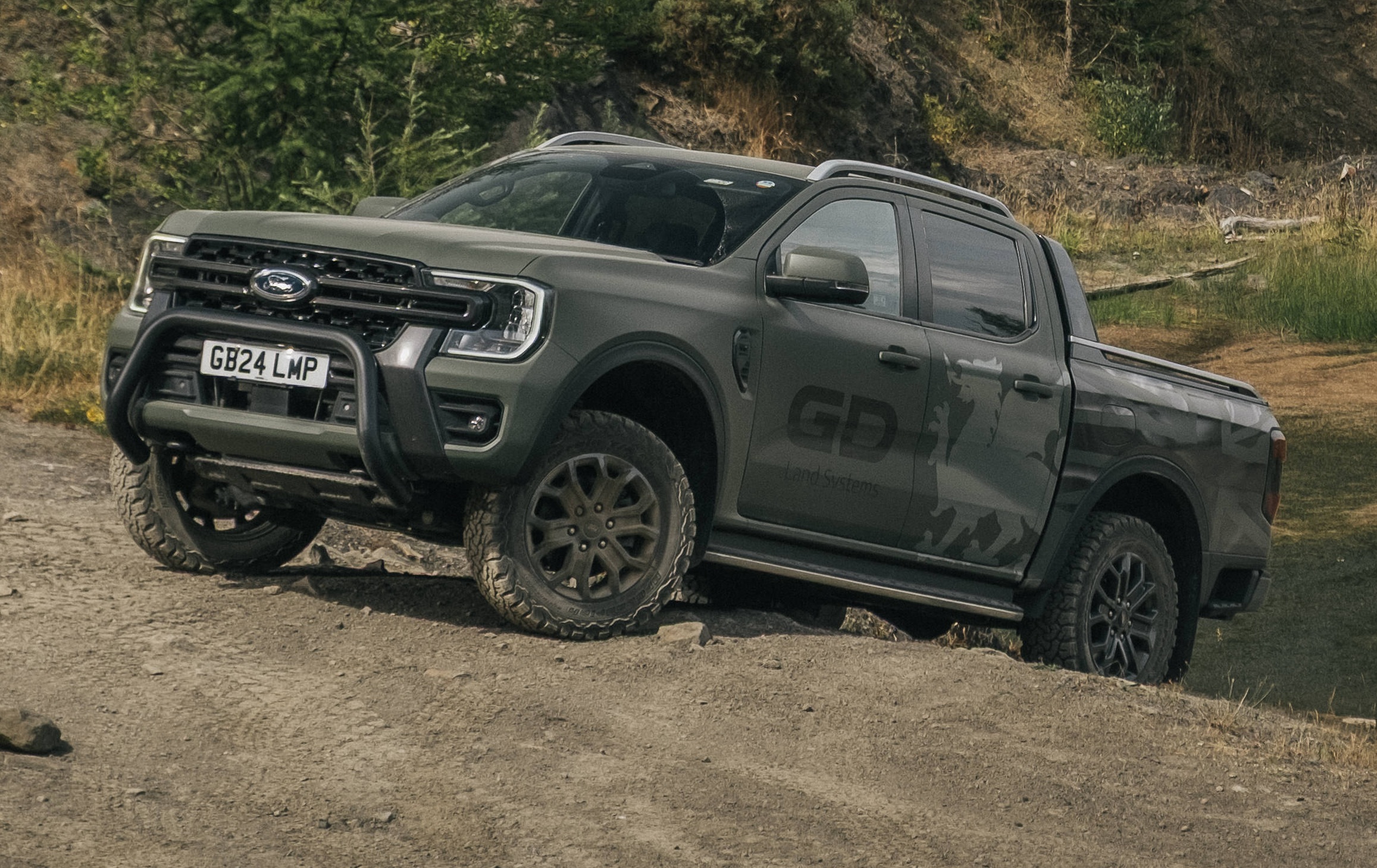 Picape Ford Ranger cinza-escura com para-choque de proteção preto e pneus off-road, subindo uma rampa de terra com vegetação ao fundo