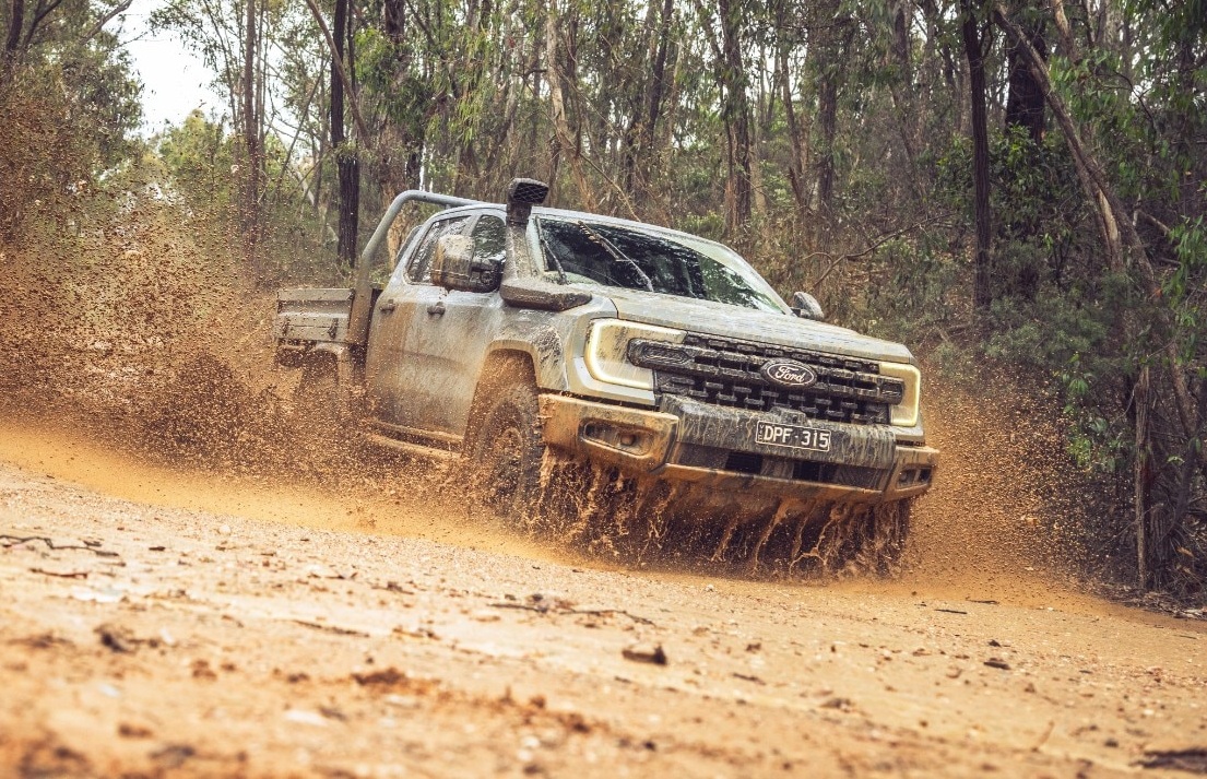 Picape Ford Ranger cinza escura, com snorkel e para-choque robusto, atravessando uma trilha de terra e lama, espalhando barro para o lado esquerdo. O veículo está sujo de lama, em meio a uma floresta densa com árvores altas