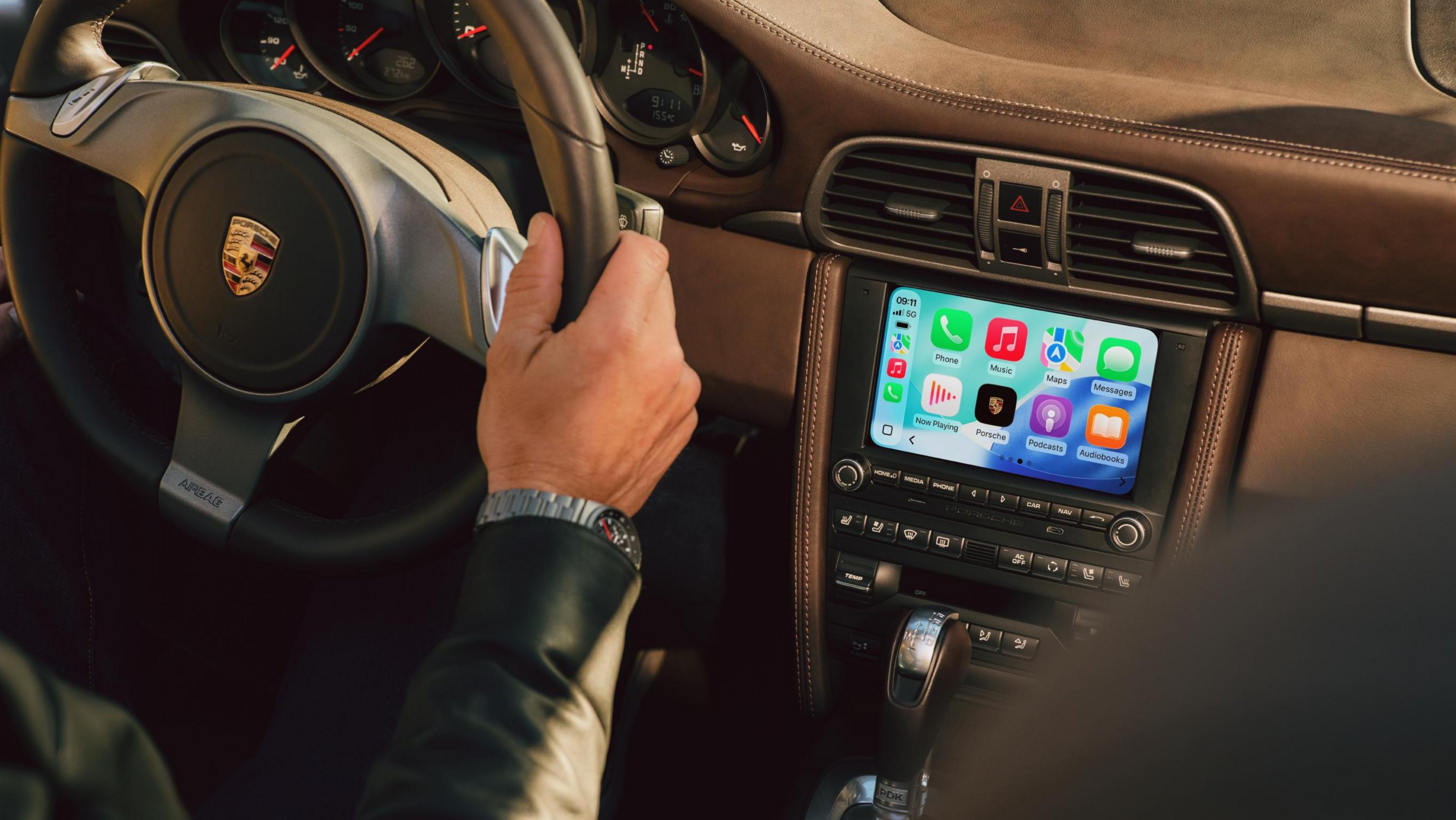 Mão de uma pessoa segurando o volante de um carro Porsche, com o painel de instrumentos e a tela do sistema de infoentretenimento exibindo o CarPlay com ícones de aplicativos