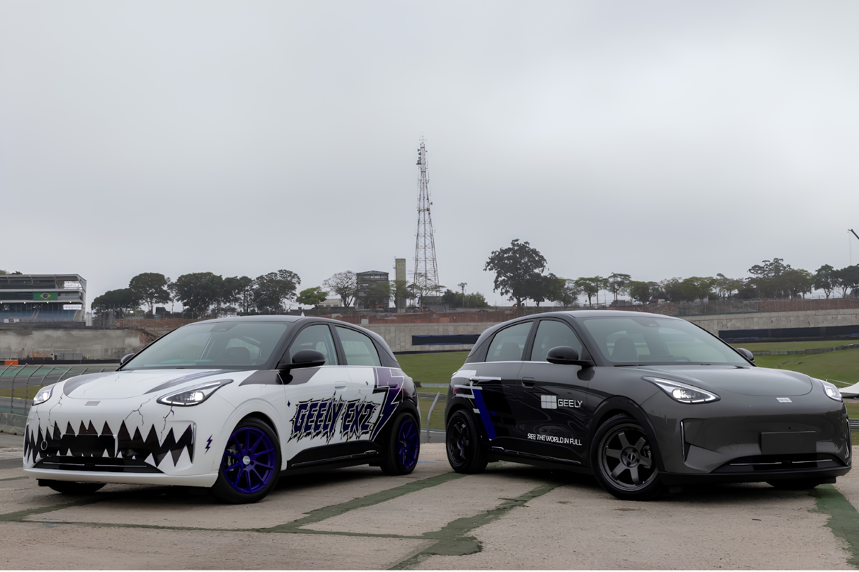Dois carros elétricos Geely EX3, um branco com grafismos de dentes e rodas azuis, e outro cinza escuro com detalhes azuis, estacionados em uma pista de corrida sob céu nublado. Ao fundo, árvores e uma torre de transmissão.