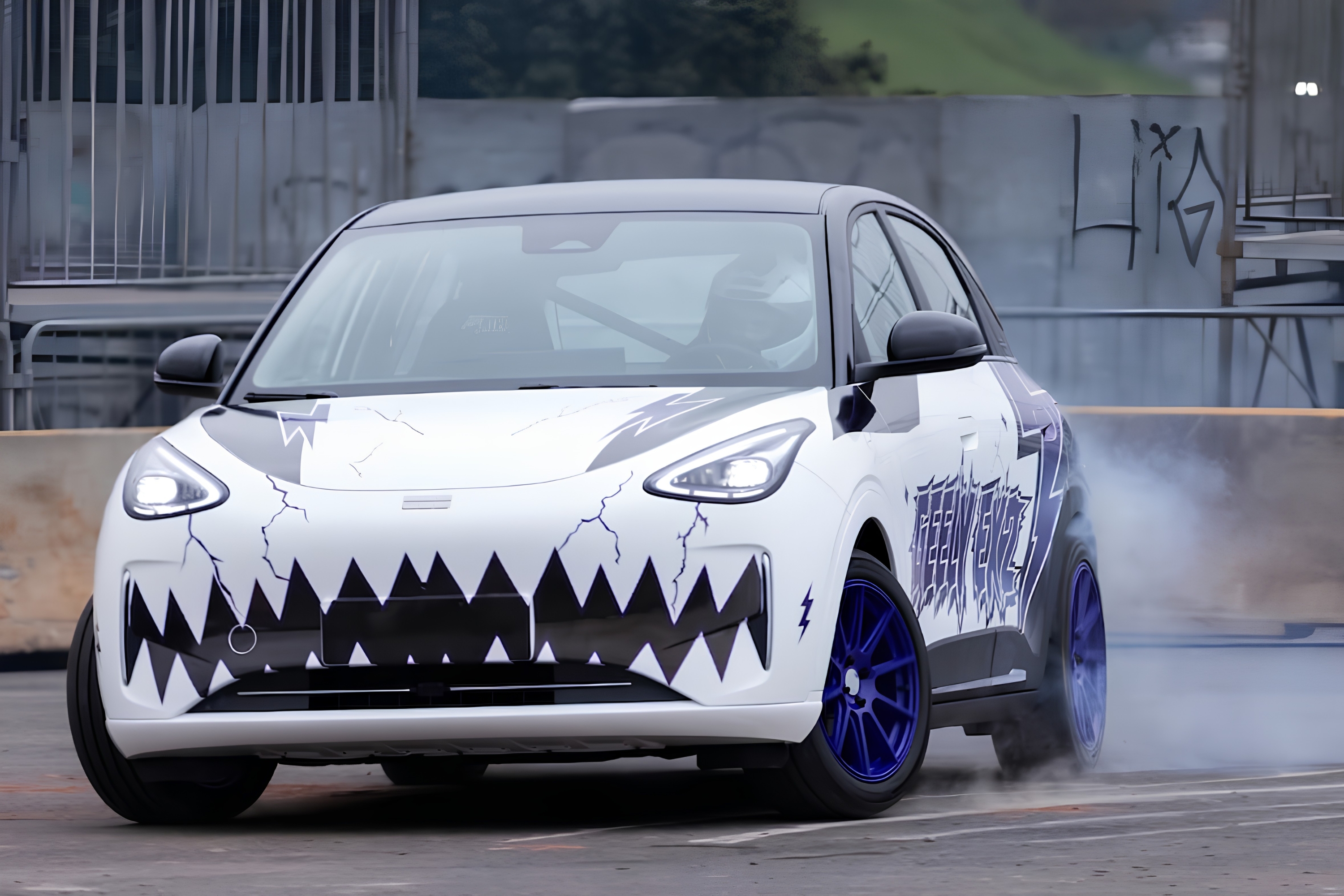 Carro branco com teto preto e rodas azuis, adesivado com boca de tubarão e raios, fazendo drift com fumaça saindo do pneu traseiro, em pista de concreto com grades e muro ao fundo