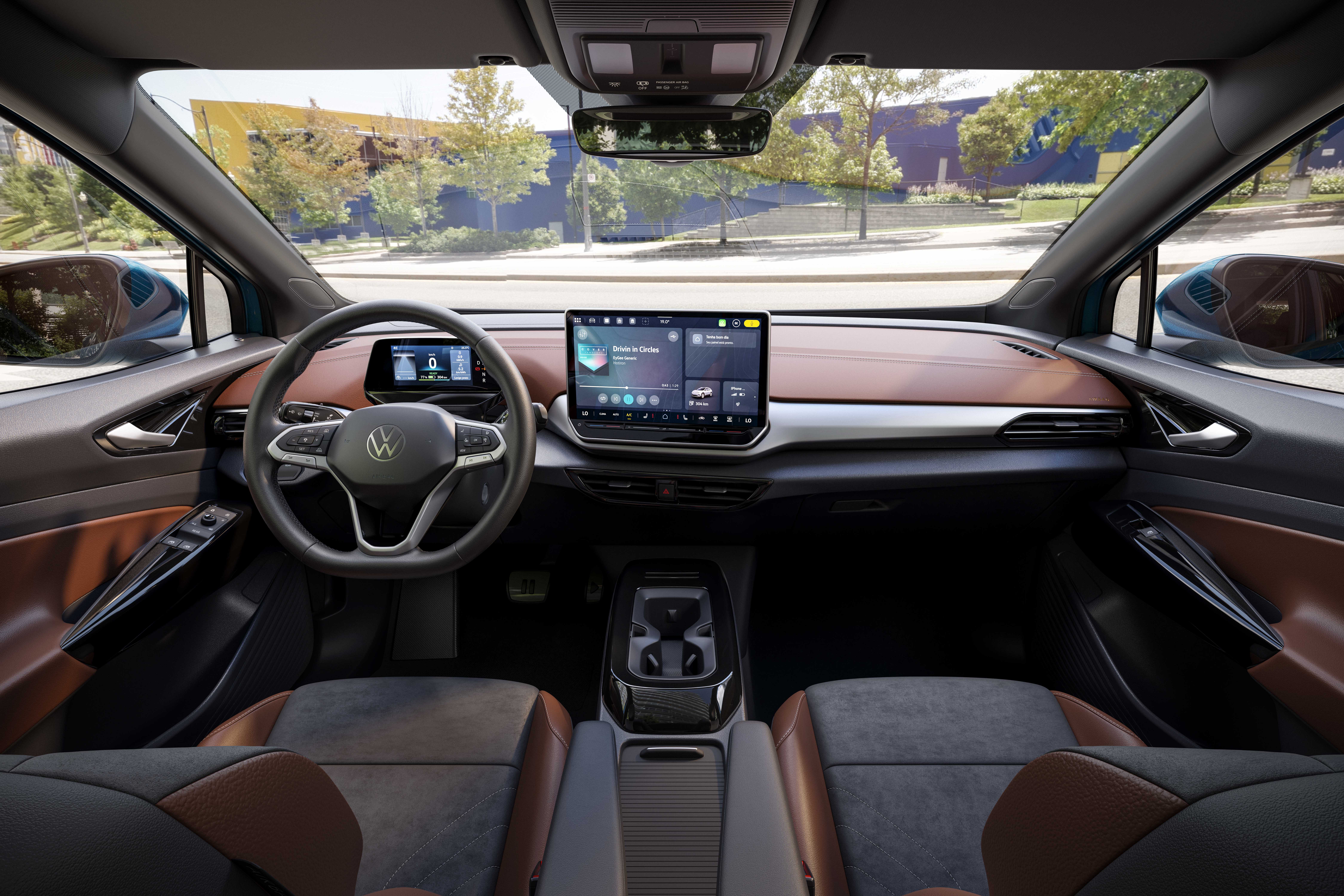 Interior de um carro moderno, com painel em tons de marrom e cinza. O volante tem o logo da Volkswagen e há duas telas digitais: uma menor atrás do volante e uma maior central, exibindo um mapa e controles de mídia. Os bancos são bicolores, cinza e marrom, e o console central possui porta-copos. A visão através do para-brisa mostra uma rua arborizada e um prédio azul ao fundo