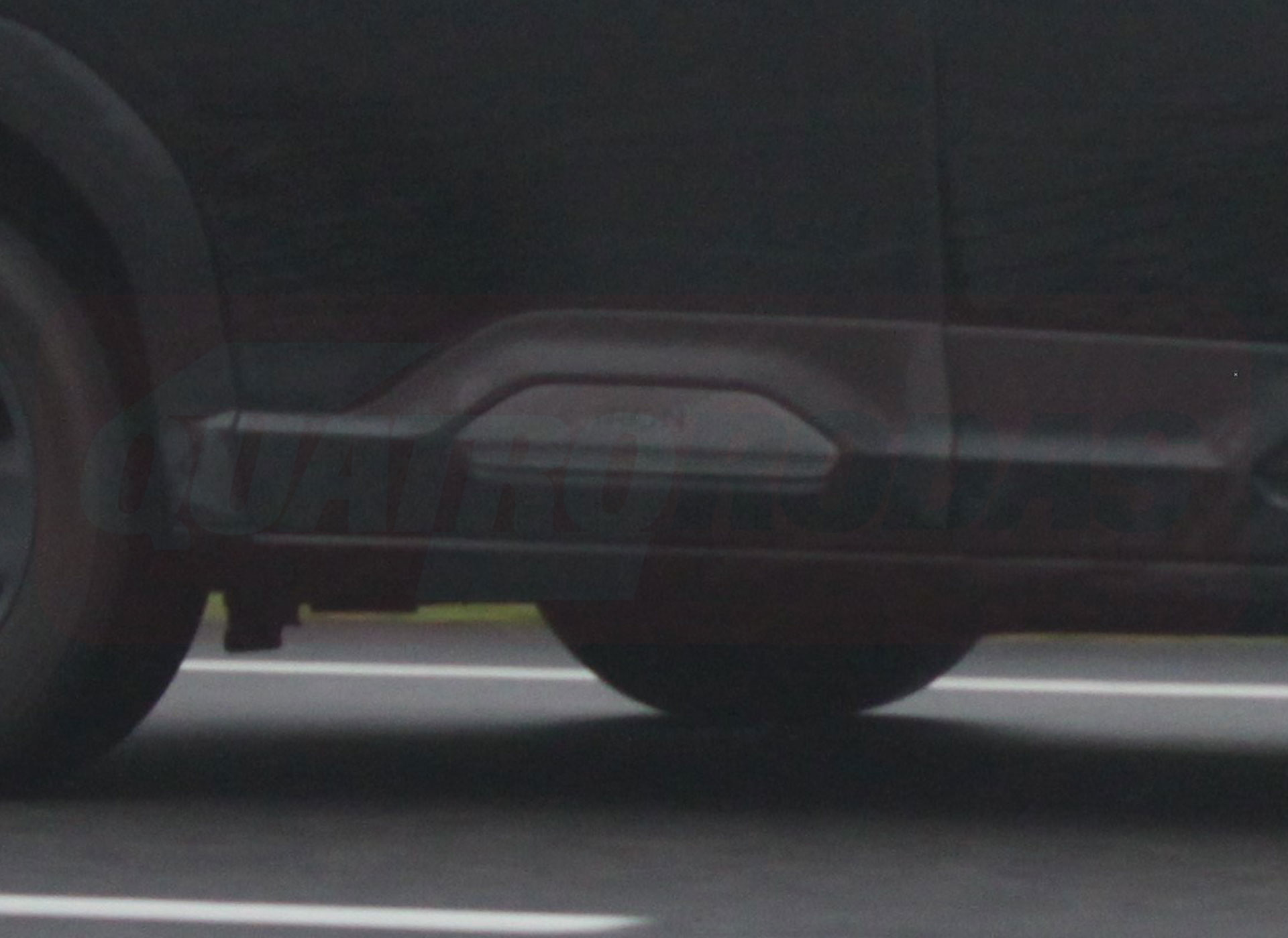 Lateral de um carro escuro, possivelmente preto ou cinza chumbo, com detalhes de design na parte inferior da porta e um pneu visível à esquerda. O veículo está sobre uma superfície cinza escura, que parece ser asfalto, com duas linhas brancas paralelas marcando o chão. Há um reflexo avermelhado na lataria do carro