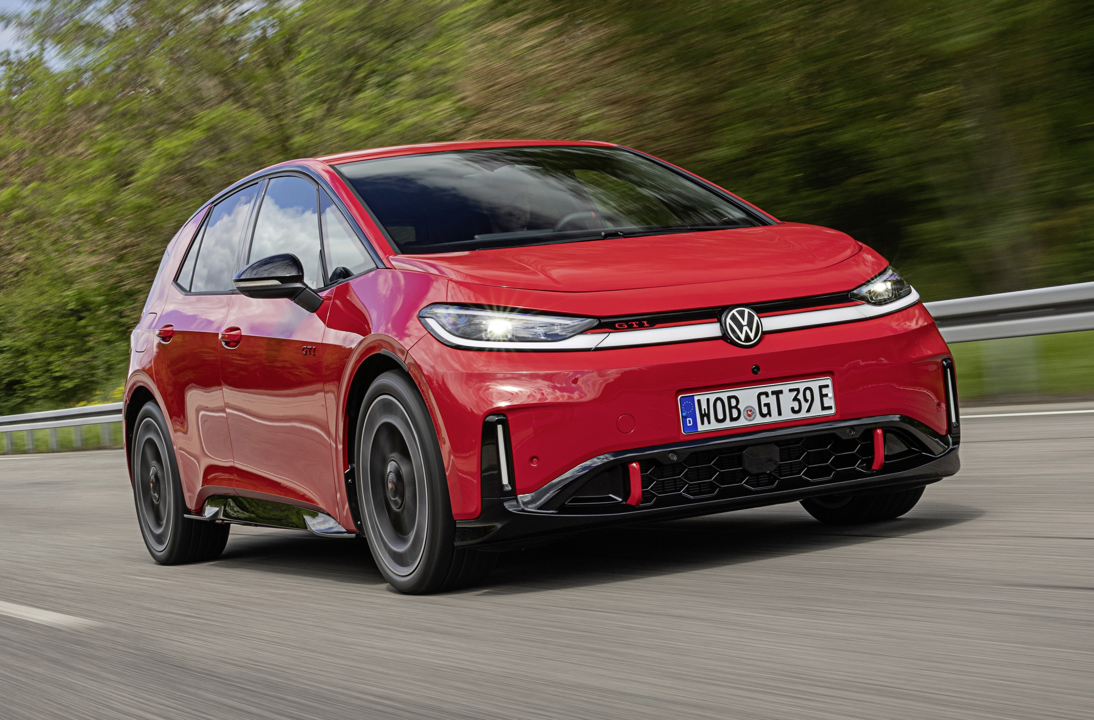 Carro vermelho Volkswagen ID.3 GTI em movimento, visto de frente e levemente de lado, em uma estrada com vegetação borrada ao fundo. O veículo tem faróis acesos, grade frontal preta com detalhes vermelhos e placa alemã WOB GT 39E