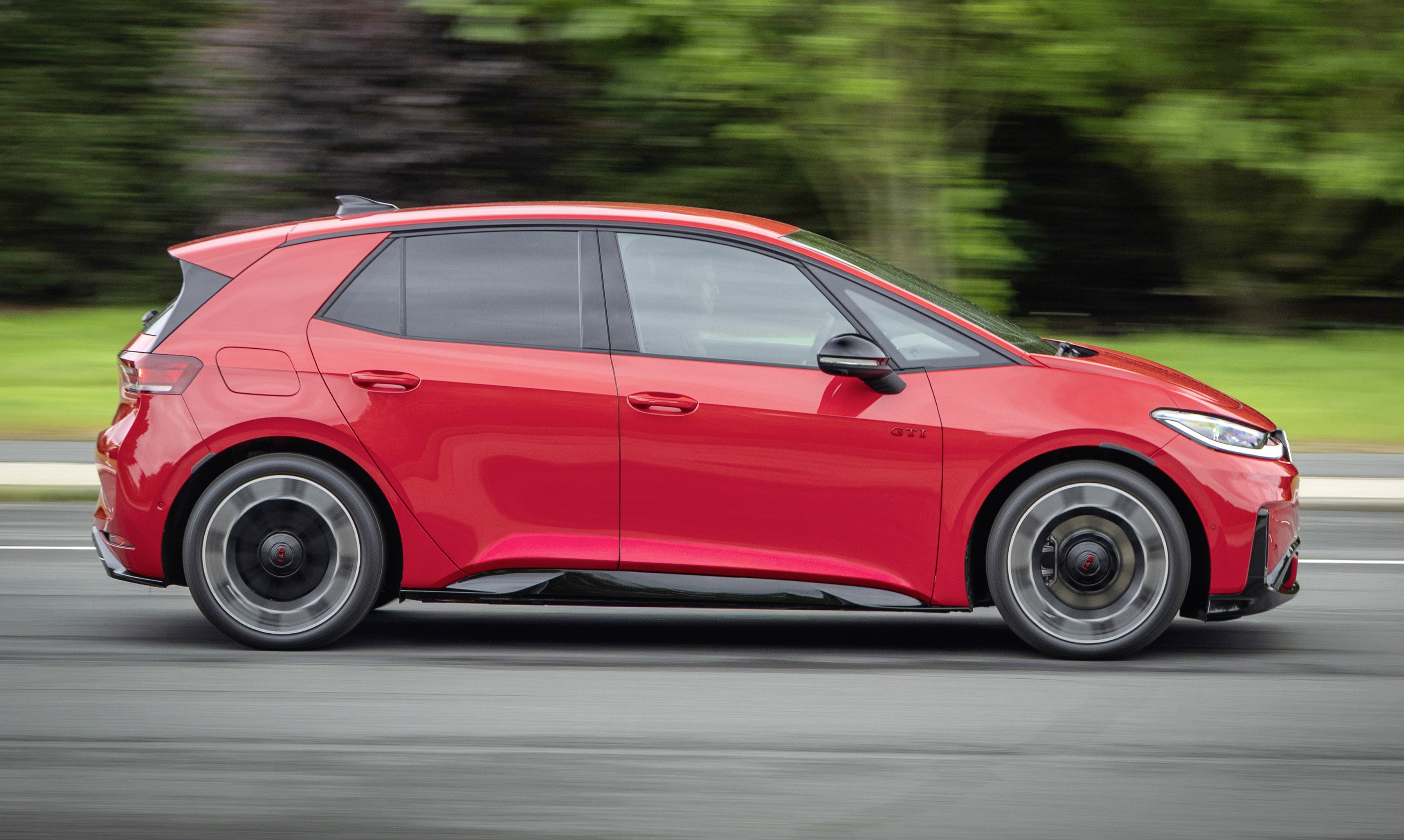 Carro compacto vermelho em movimento, visto de lado, com rodas girando rapidamente e fundo borrado de árvores verdes