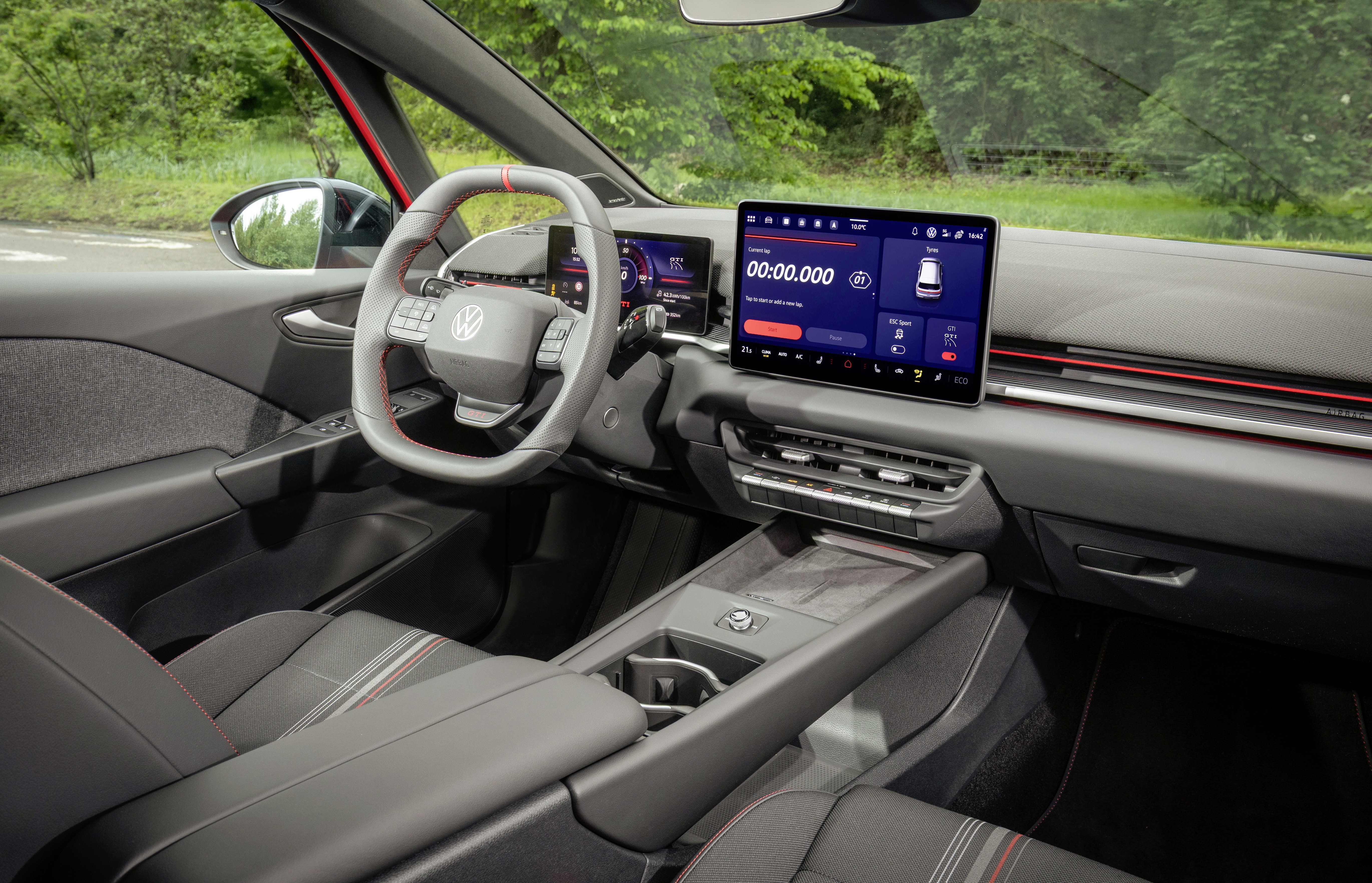 Interior de um carro moderno, com volante cinza e detalhes vermelhos, painel digital e uma tela multimídia grande exibindo um cronômetro e informações do veículo. O banco do motorista é cinza escuro com listras vermelhas e brancas. A paisagem verde de árvores é visível através do para-brisa e da janela lateral