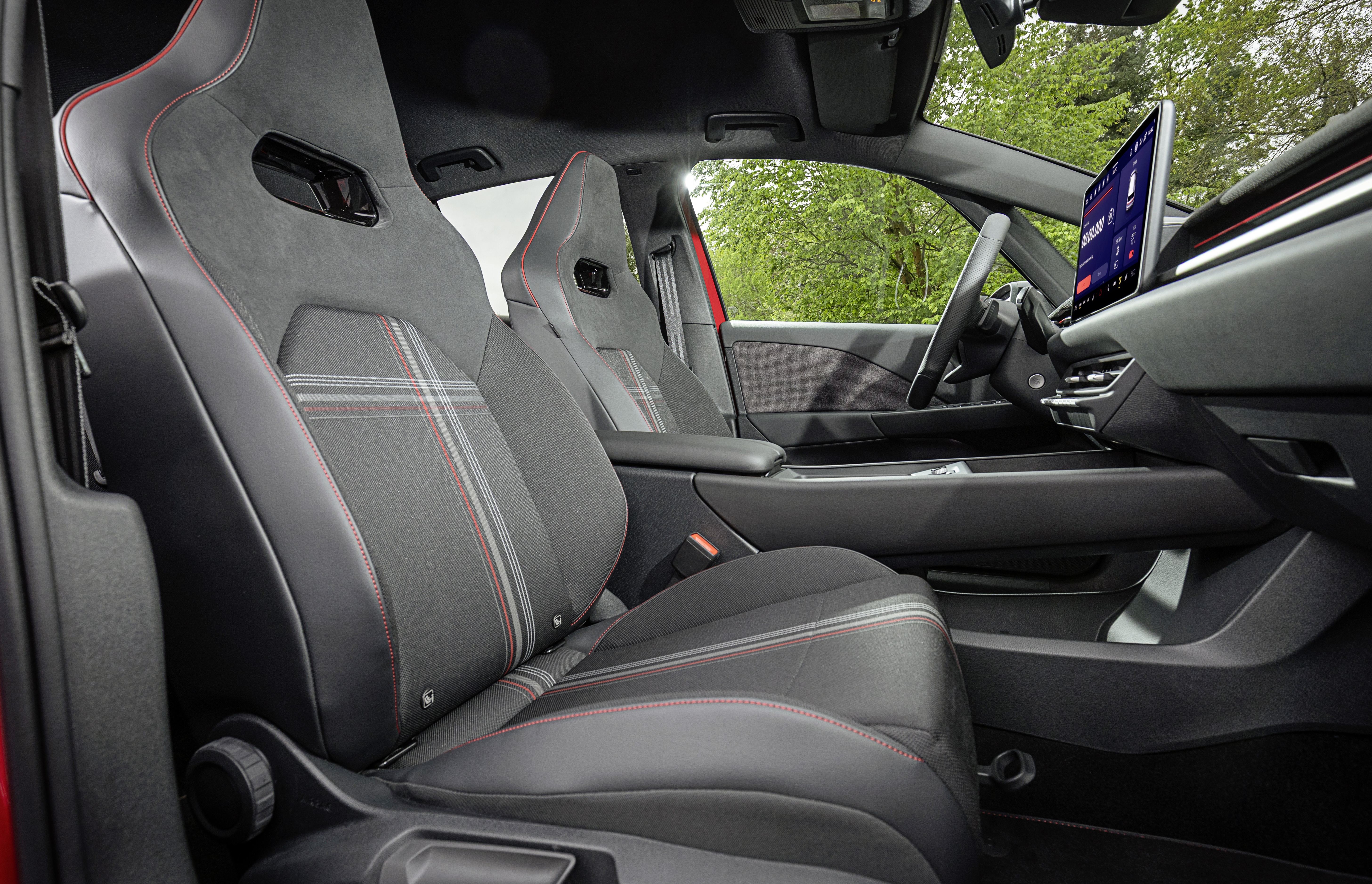 Interior de carro com bancos esportivos cinza-escuros e detalhes em vermelho, costuras vermelhas e padrão xadrez. Volante e painel pretos, tela multimídia vertical no centro. Vista externa de árvores verdes pela janela.