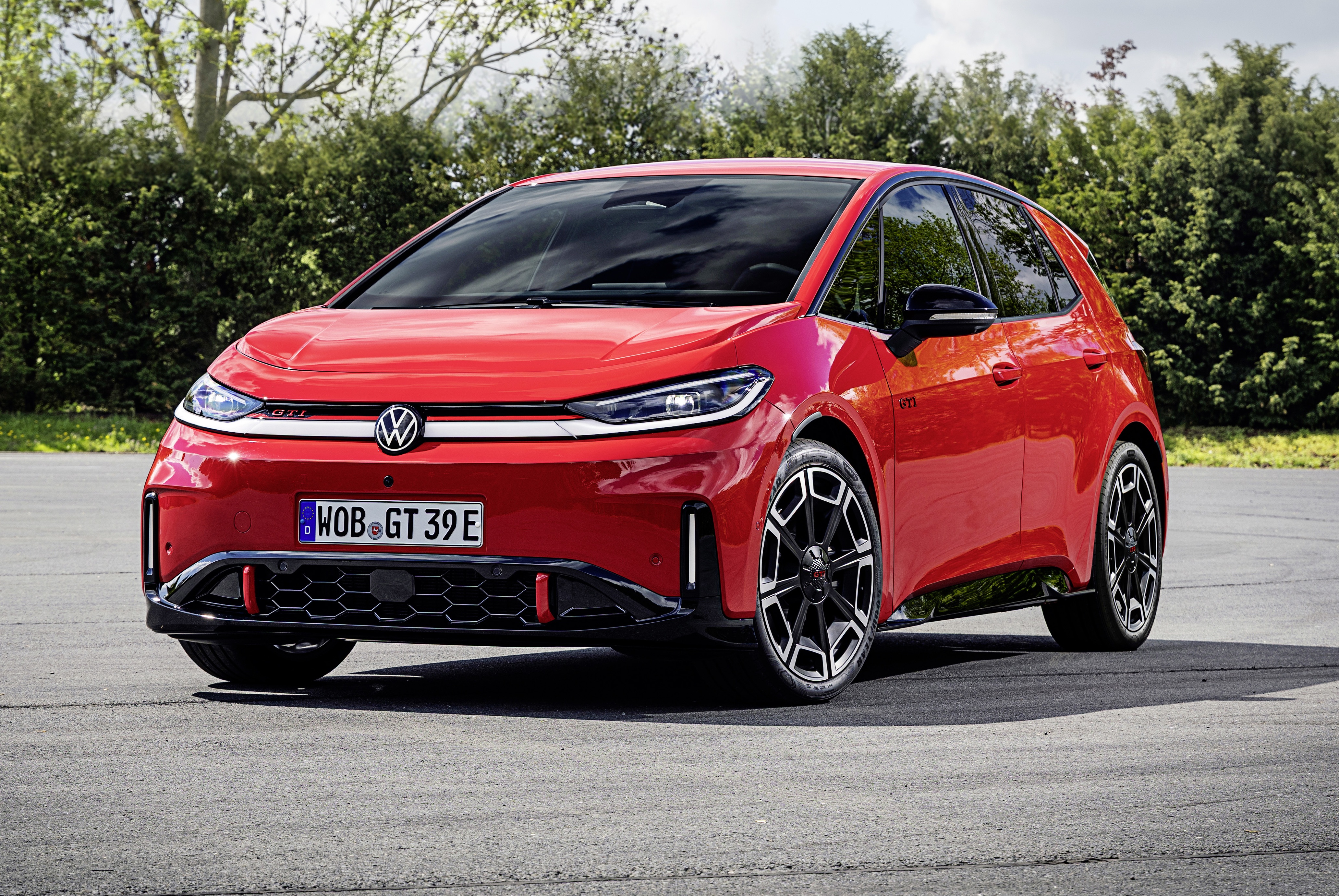 Carro elétrico Volkswagen ID.3 vermelho vibrante, com detalhes em preto e rodas esportivas, estacionado em uma pista de asfalto com árvores e arbustos ao fundo, sob luz solar