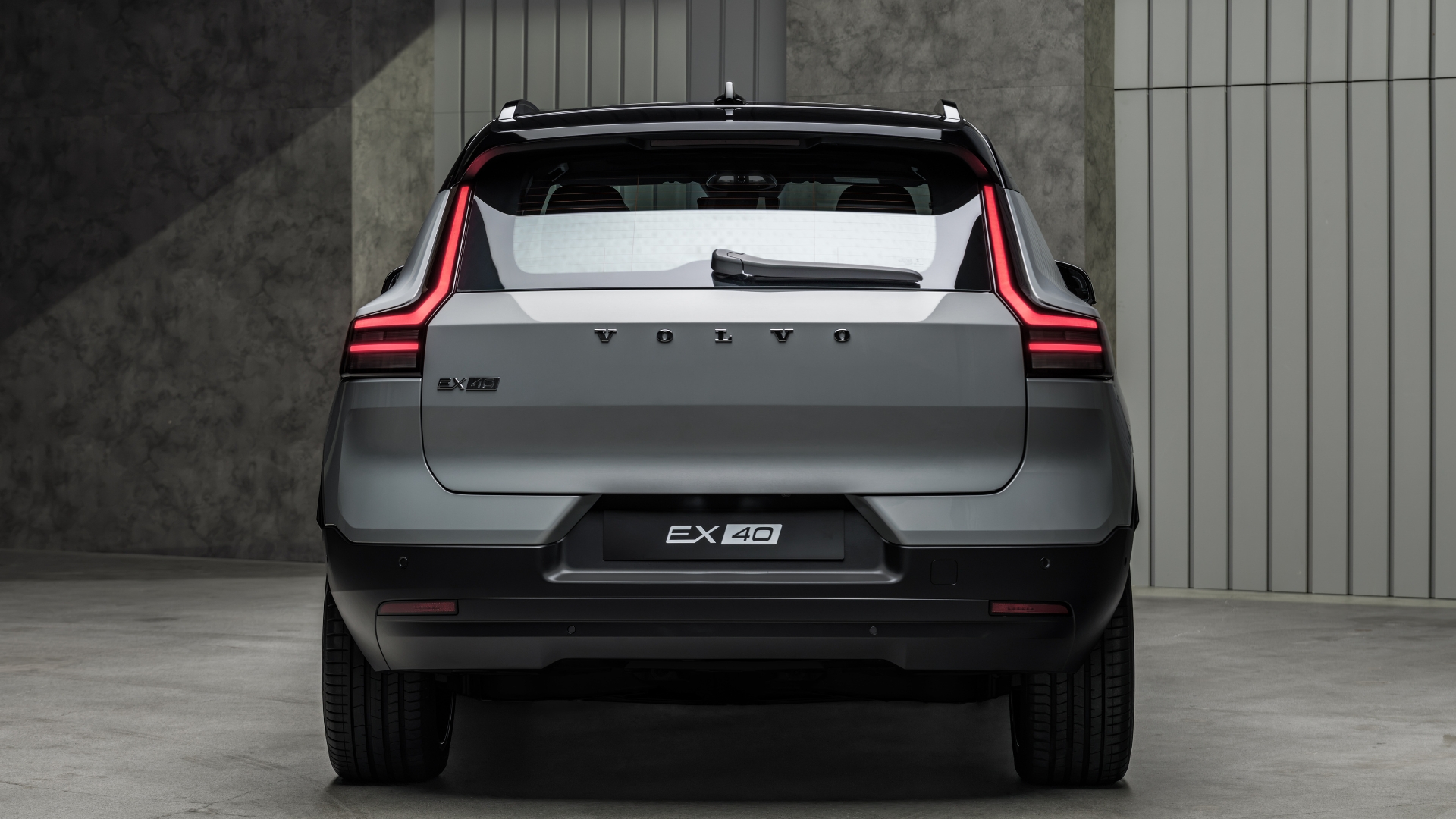 Traseira de um carro Volvo EX40 cinza-chumbo, com lanternas verticais em LED acesas, limpador de para-brisa traseiro e teto preto. O veículo está estacionado em um ambiente interno com paredes de concreto e iluminação suave
