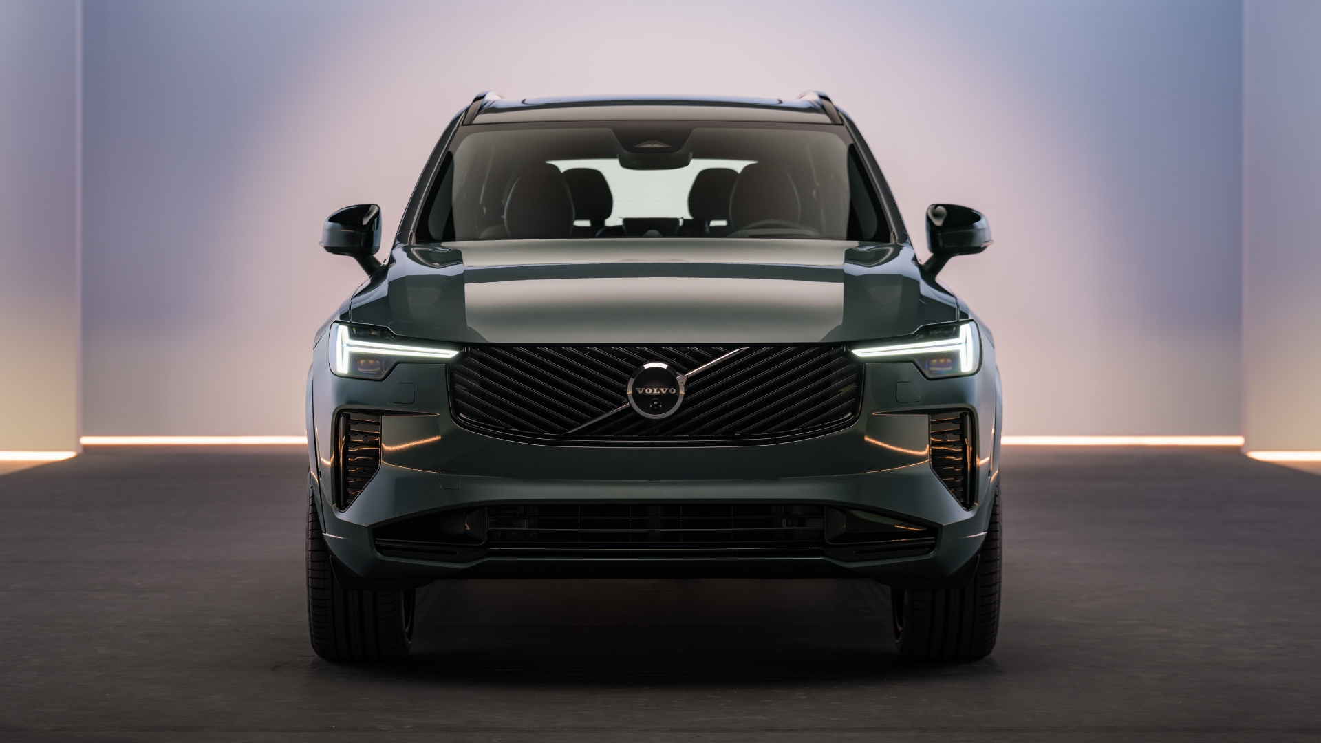 SUV Volvo escuro, verde-azulado, visto de frente, com faróis e luzes diurnas acesas em forma de T, grade frontal preta e logotipo da marca. O carro está em um ambiente interno com paredes claras e uma linha de luz horizontal na base