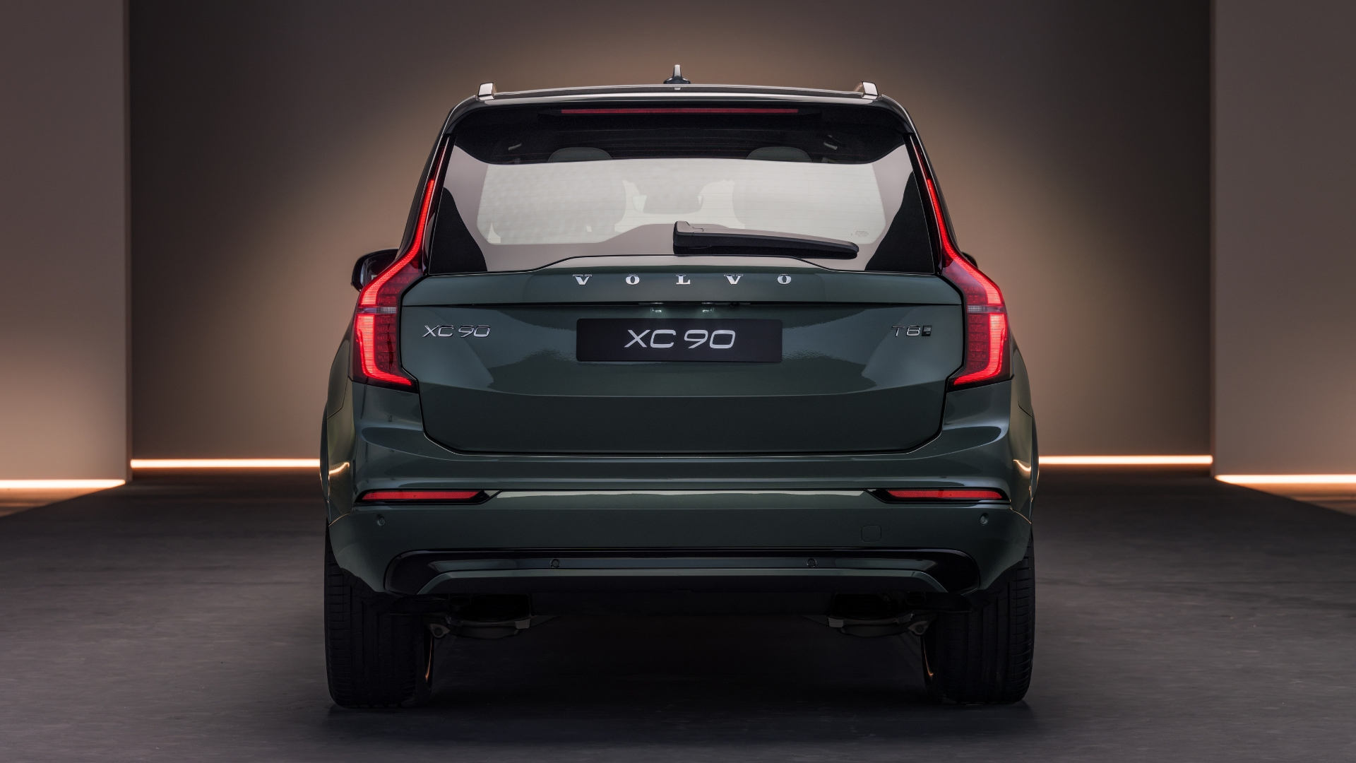 Traseira de um SUV Volvo XC90 verde escuro, com lanternas verticais acesas e o nome do modelo em destaque na tampa do porta-malas, em um ambiente escuro com iluminação horizontal no chão