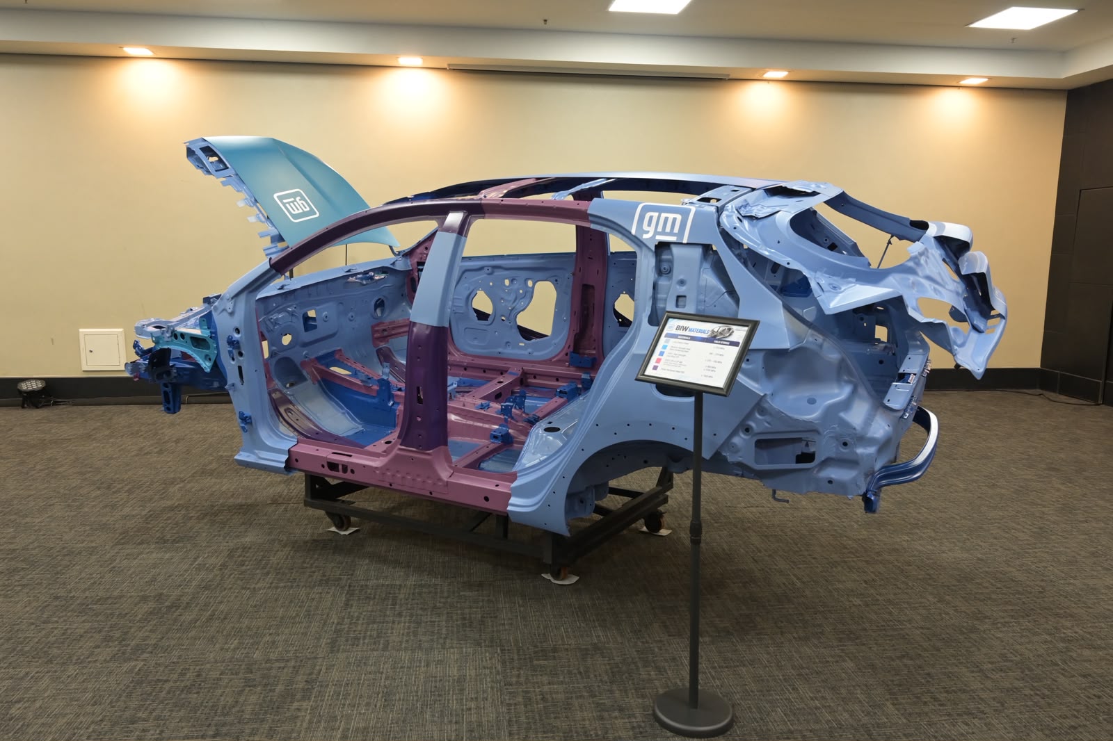 Carroceria de veículo em exposição, com estrutura metálica azul e roxa, sem portas ou partes internas, sobre um suporte preto. Um painel informativo está à direita, e o logotipo GM aparece na lateral e no capô aberto
