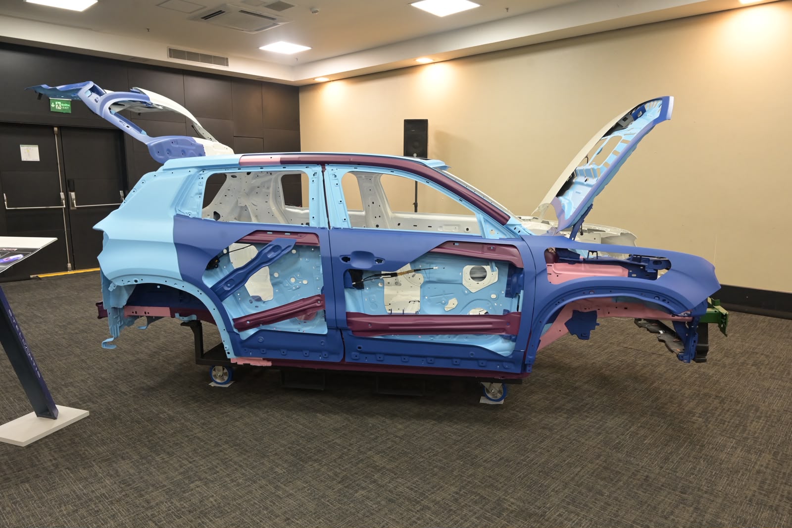 Carroceria de um veículo em exposição, com partes em azul claro, azul escuro, rosa e branco, mostrando a estrutura interna. O capô e a tampa traseira estão abertos, revelando o esqueleto do carro. O veículo está sobre um piso de carpete cinza, em um ambiente interno com paredes claras e escuras, e iluminação no teto