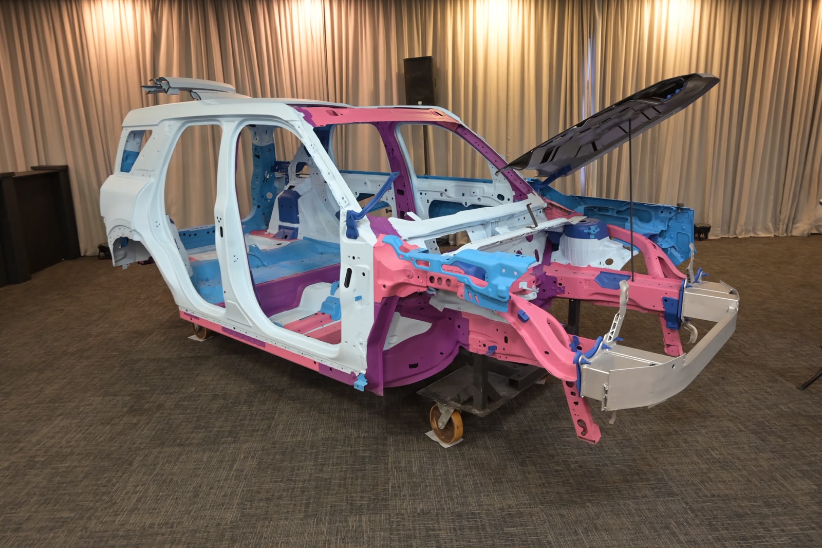 Carroceria de um veículo em construção, com partes da estrutura pintadas em branco, rosa, azul e roxo, destacando os diferentes materiais e componentes. O capô está aberto, revelando a parte interna do motor. O carro está sobre um suporte em um ambiente interno com cortinas bege ao fundo e carpete marrom no chão