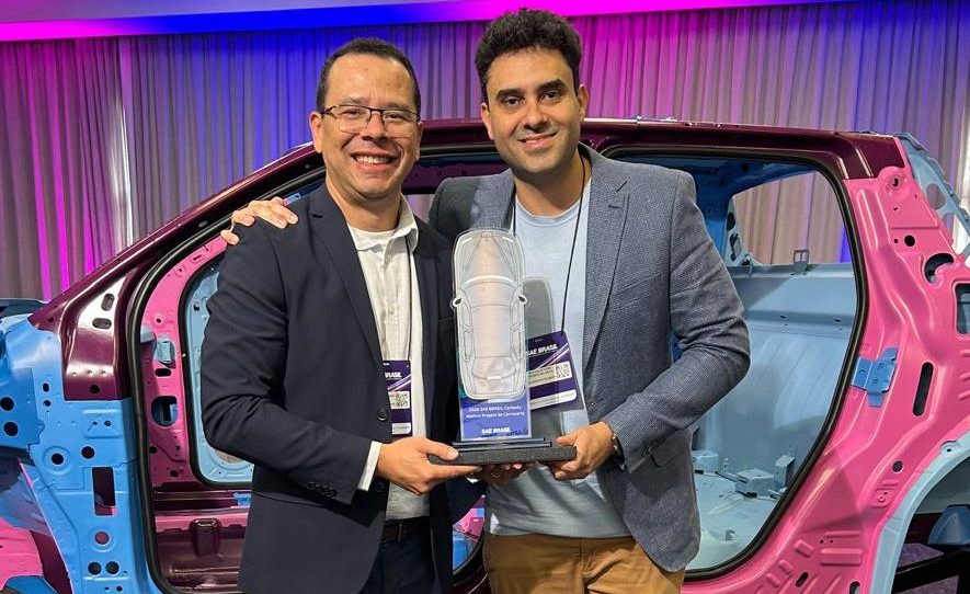 Dois homens sorridentes, um de óculos e blazer azul-marinho, outro de blazer cinza, seguram um troféu transparente em formato de carro. Ao fundo, uma estrutura de carroceria de veículo em tons de rosa e azul, com iluminação roxa e azul no ambiente
