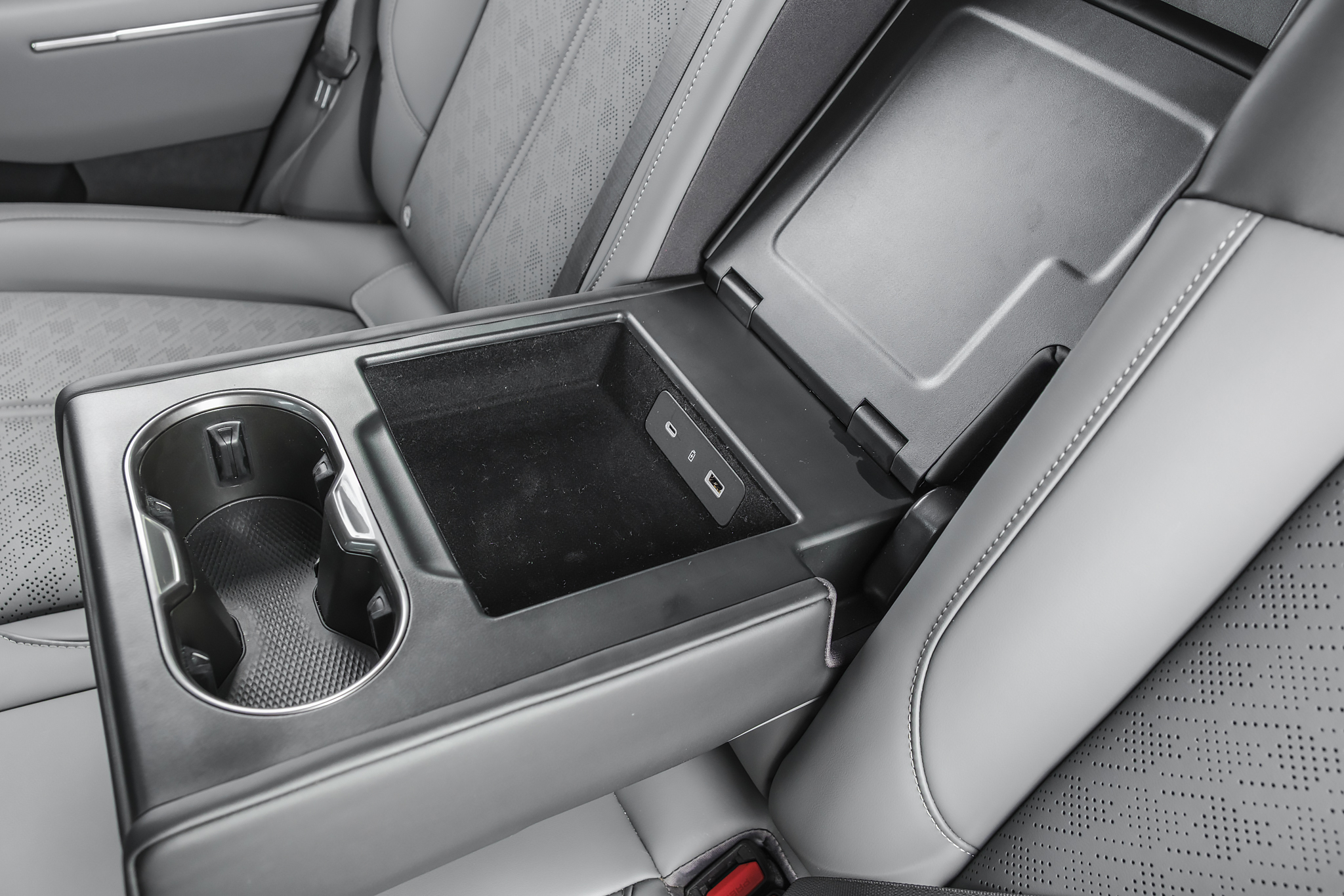 Apoio de braço traseiro de carro cinza, com porta-copos duplo e compartimento de armazenamento aberto revelando portas USB e auxiliar