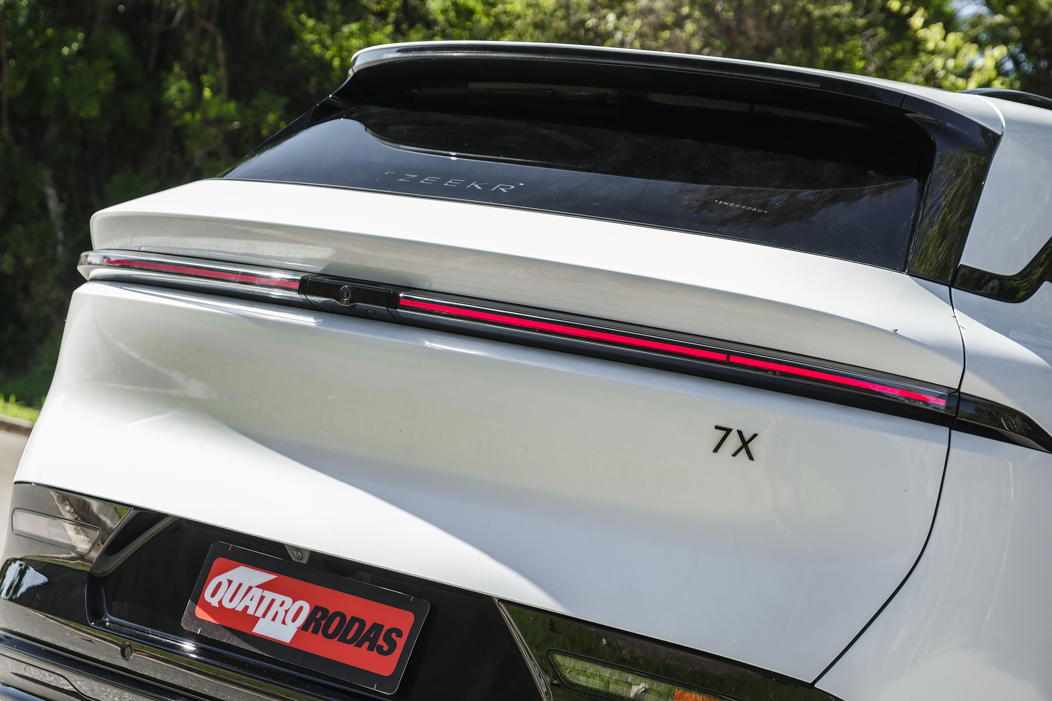 Traseira de um carro branco com lanterna vermelha horizontal, o nome ZEEKR no vidro traseiro e 7X na lataria. Abaixo, uma placa vermelha com QUATRO RODAS em branco