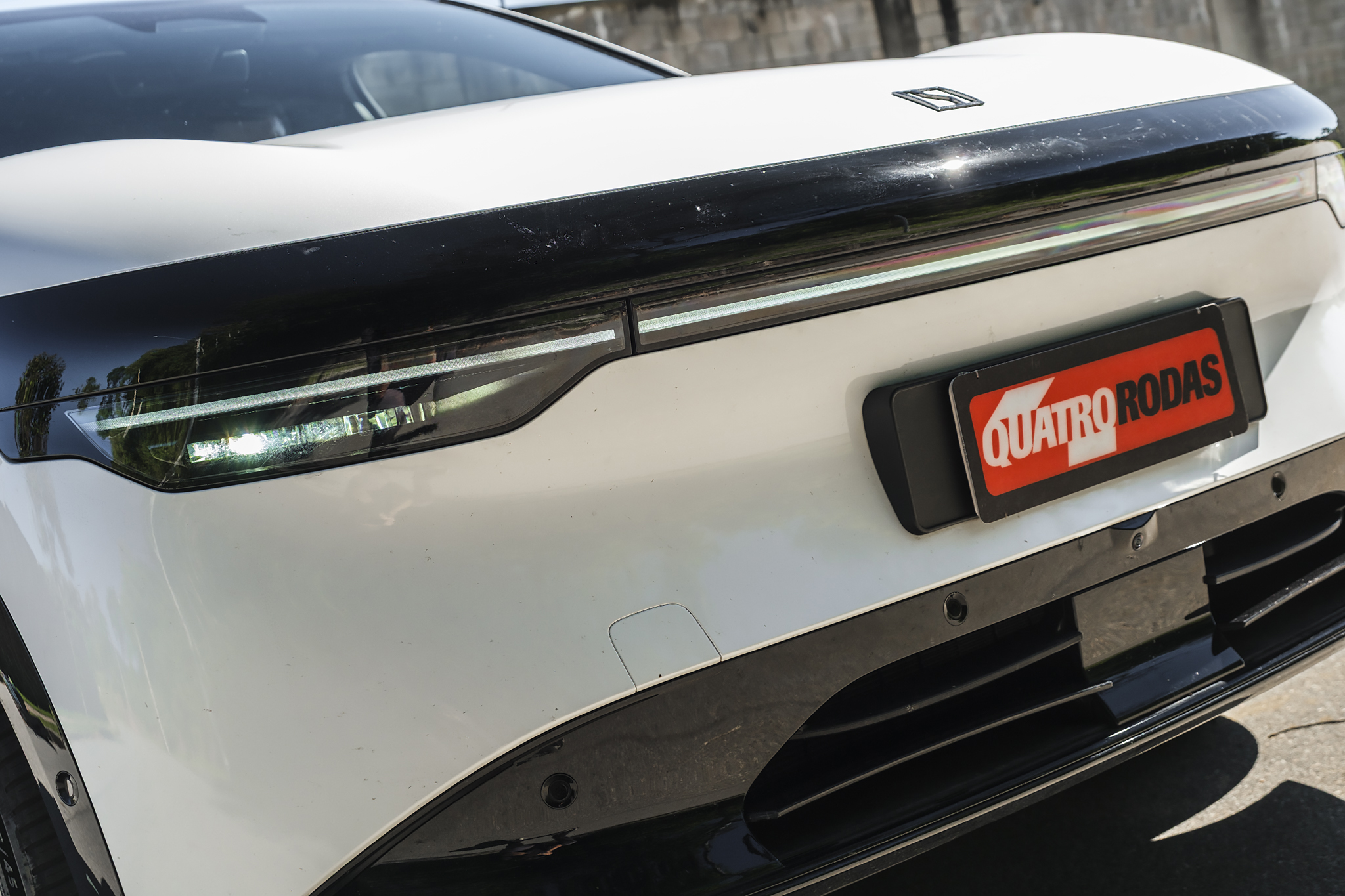 Traseira de um carro branco com uma faixa preta brilhante no porta-malas. A lanterna esquerda está acesa, e uma placa vermelha com o texto QUATRO RODAS em branco está à direita. O para-choque inferior é preto com detalhes em grade.