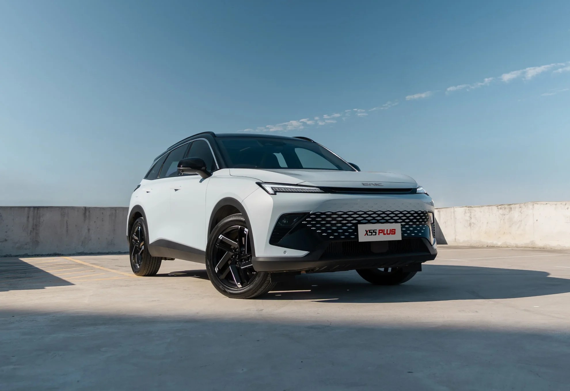 SUV branco BAIC X55 Plus, com teto preto e rodas escuras, estacionado em um piso de concreto cinza claro sob céu azul com poucas nuvens