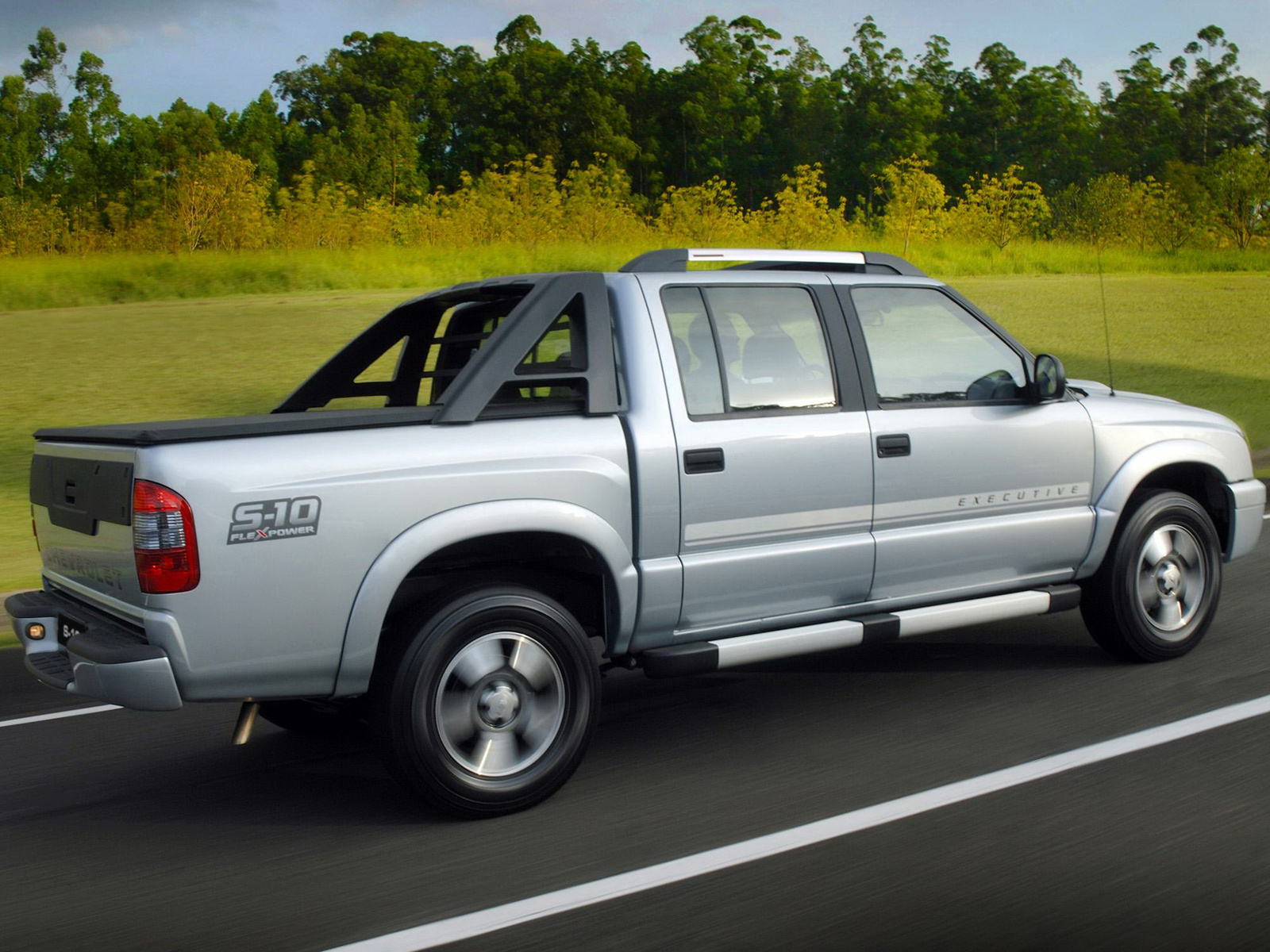 Picape Chevrolet S-10 Executive prata em movimento, vista de traseira lateral, com rodas girando. Ao fundo, vegetação verde e amarela sob céu azul
