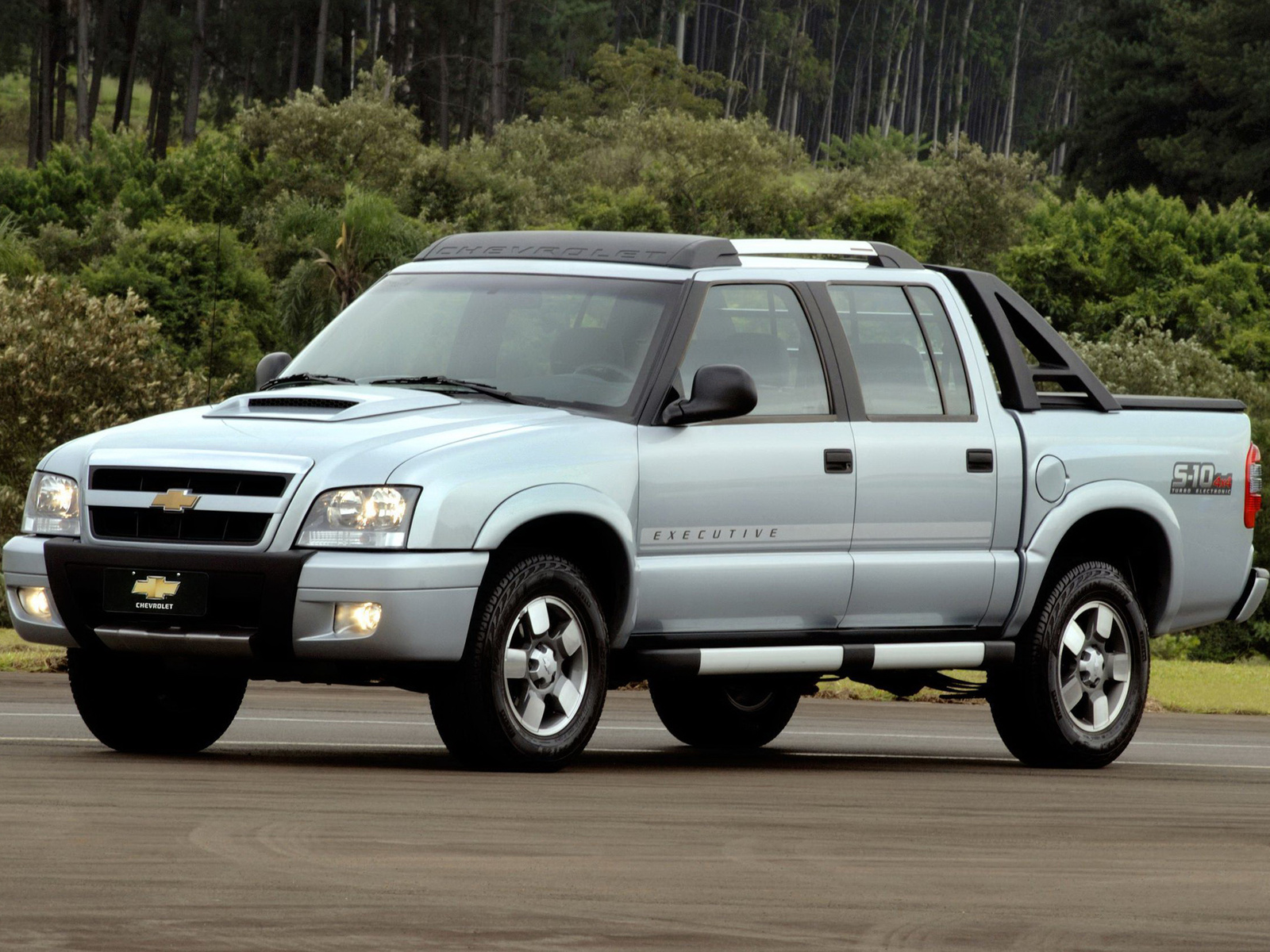 Picape Chevrolet S10 Executive prata, com cabine dupla, estribos laterais e santantônio preto, estacionada em asfalto com árvores ao fundo